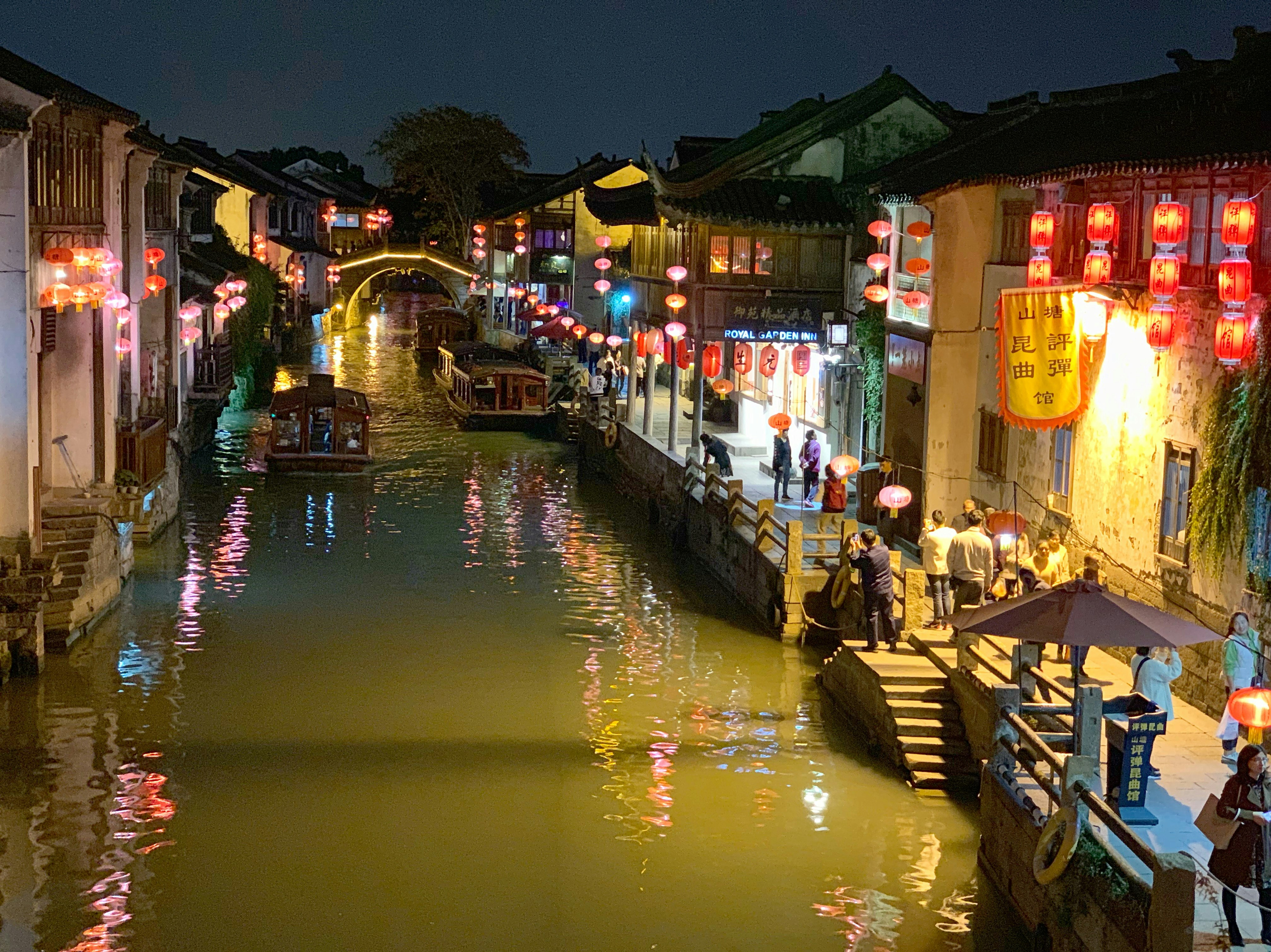 A view of one of Suzhou's canals at night. The waterway is dark while the buildings around the narrow canal are lit up by lanterns and lights. A few wooden boats travel down the canal.