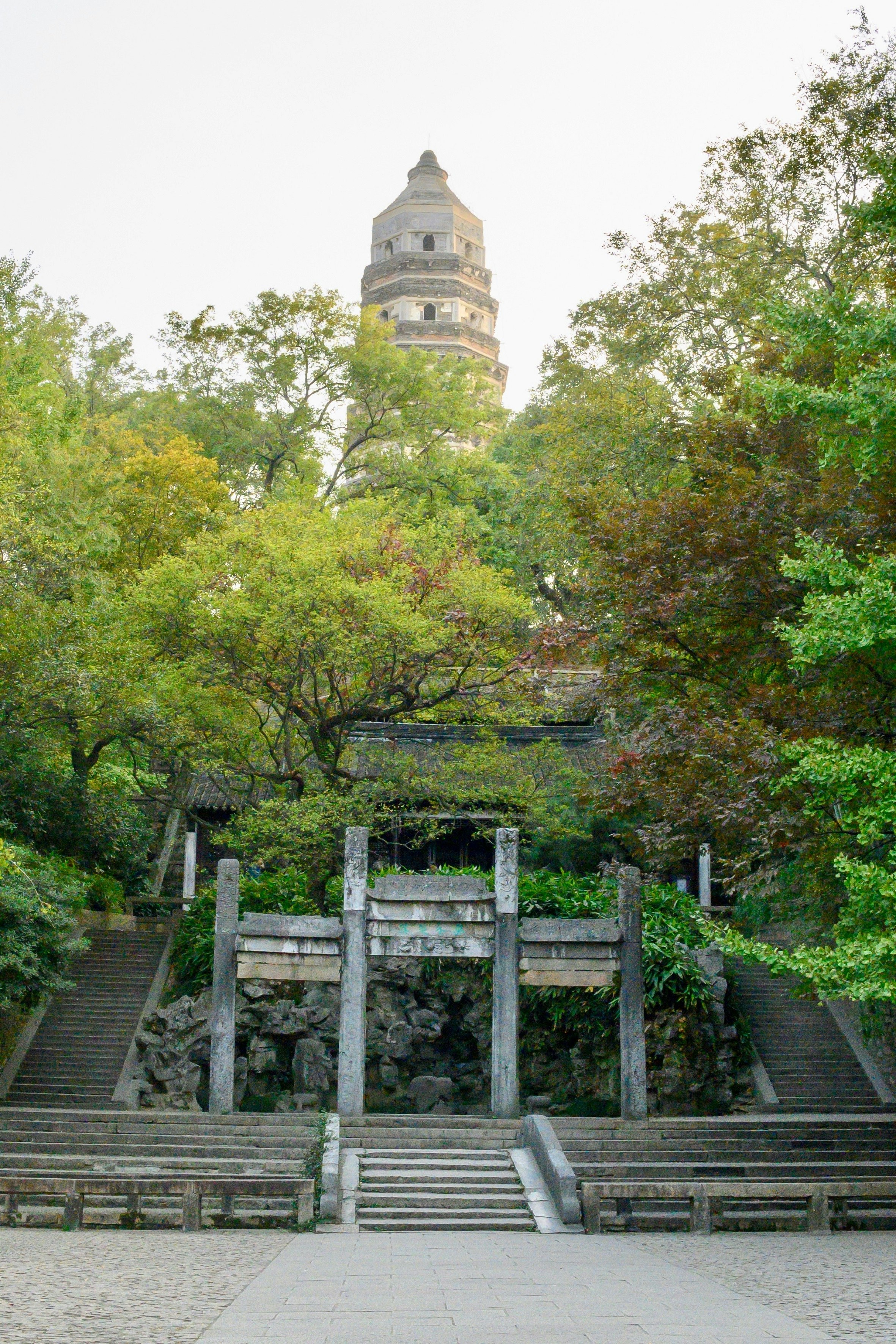 A landscape view of Tiger Hill with the brick Cloud Rock Pagoda atop it. There are steps leading up the hill, which are flanked by greenery.
