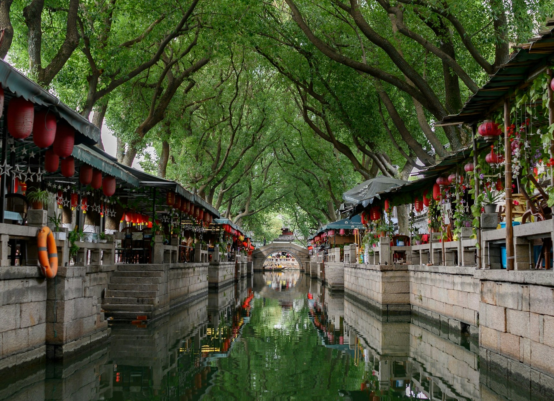 A view of a Suzhou canal, taken from the water at water level. The canal is lined on either side by small businesses, with the straight and narrow canal running under an ornate stone bridge in the distance.