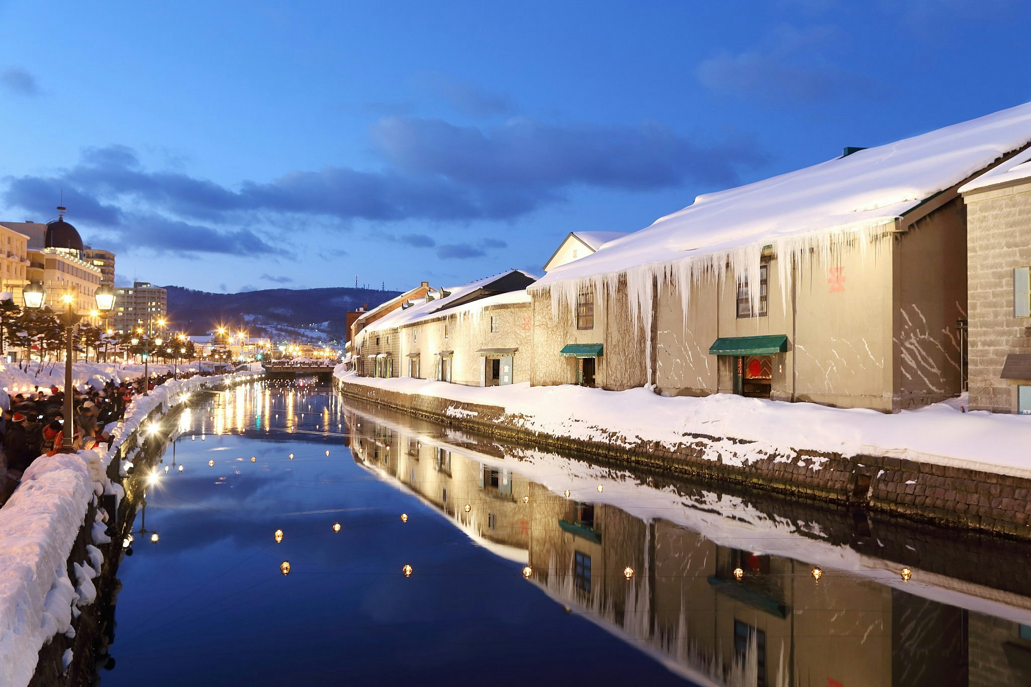 The illuminated canal of Otaru, Hokkaido, lined with snow-covered buildings, some of which are grand and imposing, at dusk.