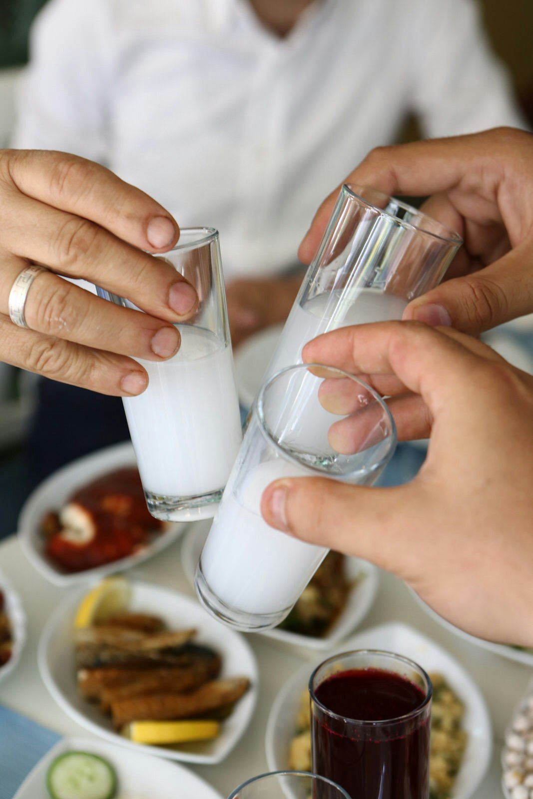 Three people clinking shot glasses of white ouzo together over a table of food