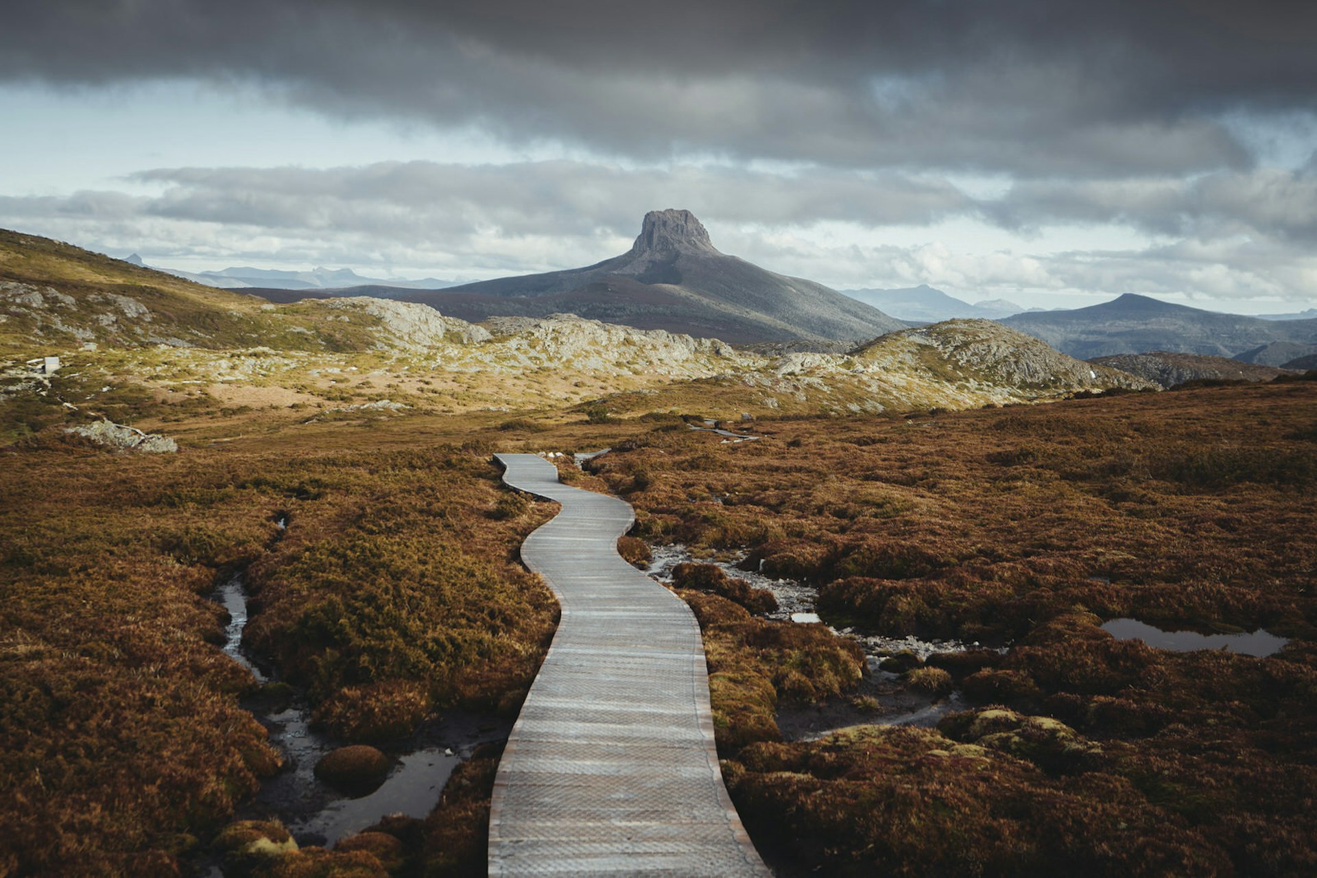 A wooden footpath stretches away from the camera over russet shrubbery and rock pools on Tasmania's Overland Trail. There is a distinctly square rocky peak in the distance underneath an overcast sky 