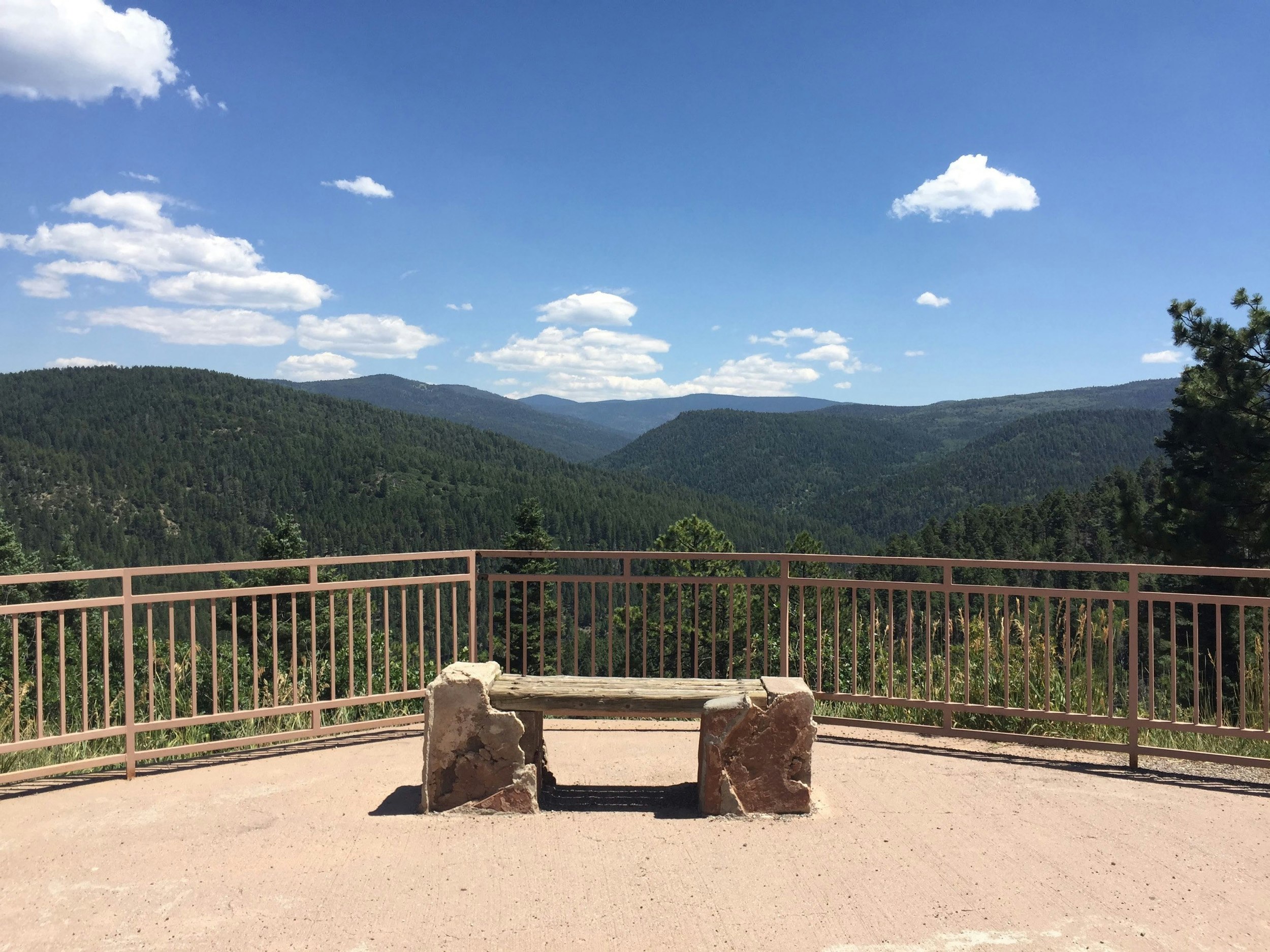 A bench is set up in front of a railing looking out over a southwestern vista on the High Road to Taos