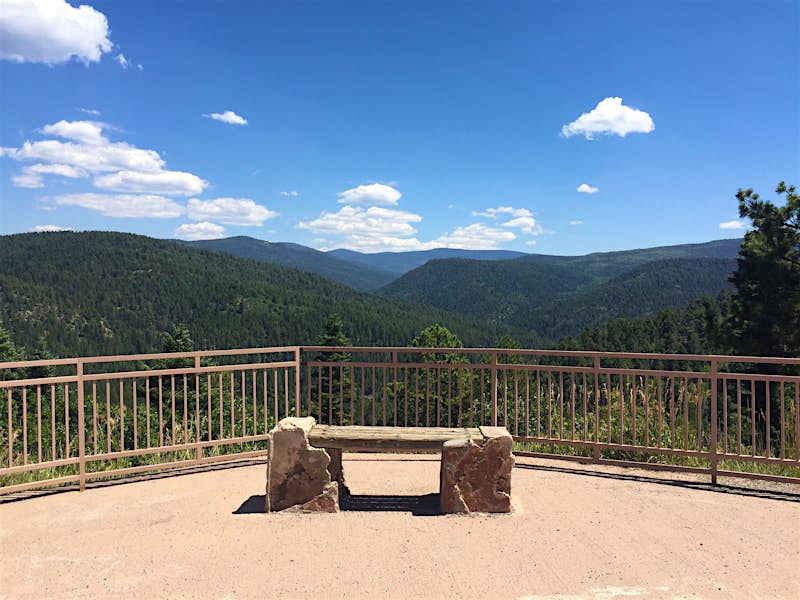 A bench is set up in front of a railing looking out over a southwestern vista on the High Road to Taos