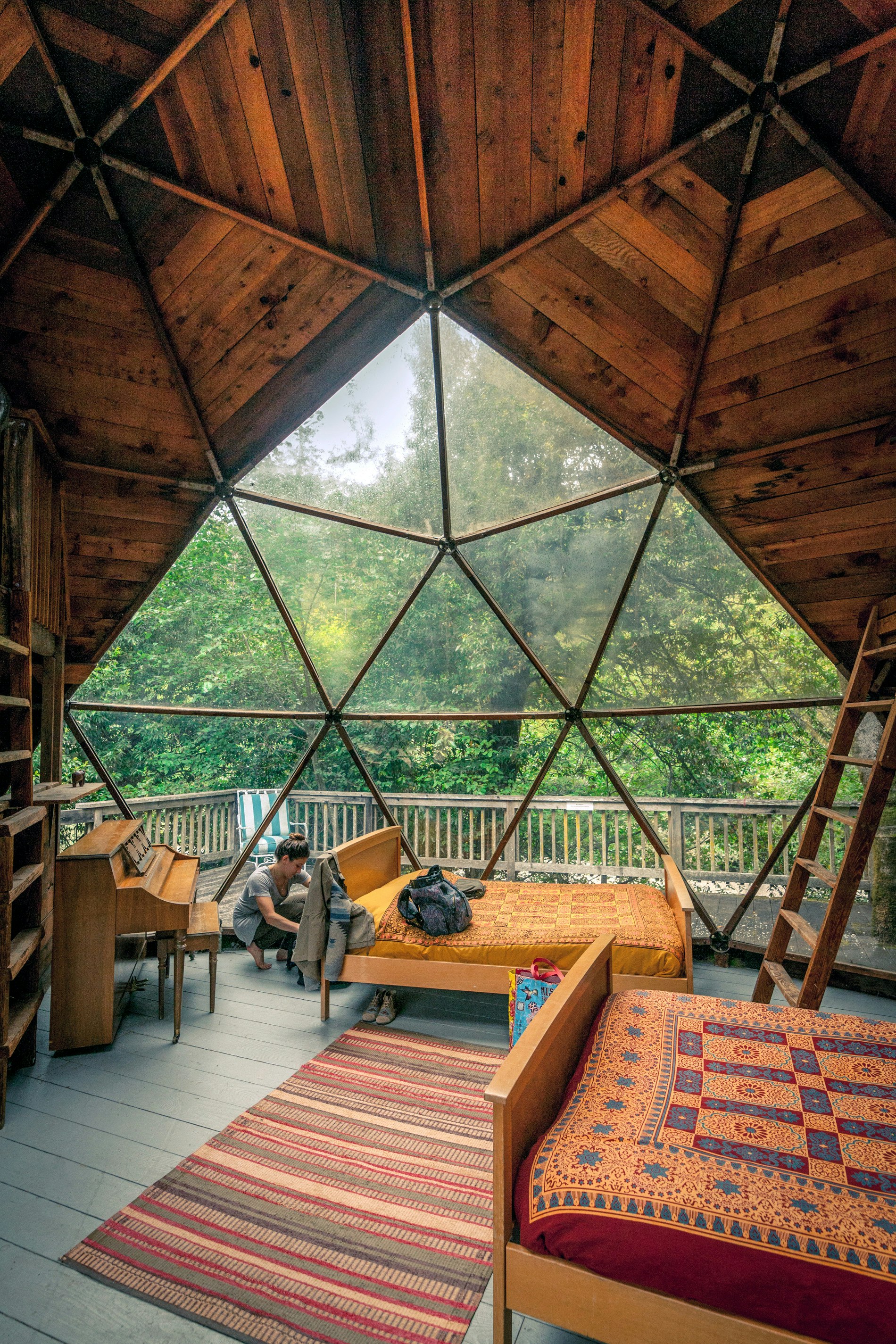 The inside of a wooden cabin. Large panes of glass make up some of the geodesic structure, creating a huge window. A woman is crouched down beside a double bed that has patchwork-printed linen. To the left is an upright piano.