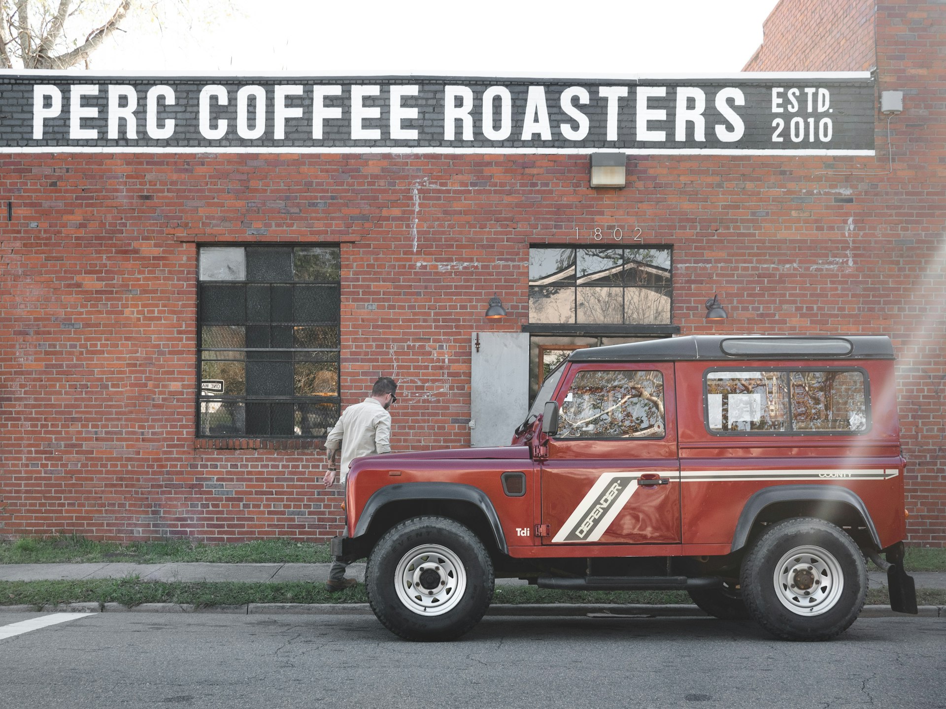 A red Land Rover Defender is parked in front of the red brick facade of Perc Coffee Roasters.