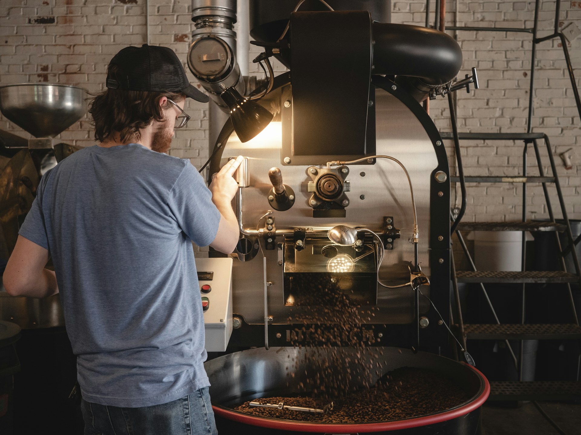 A coffee roaster with his back to the camera releases a batch of beans into a large grinding machine.