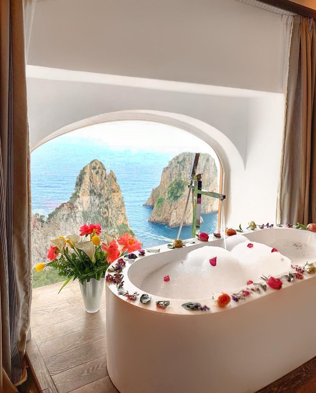 A white soaking tub filled with soap bubbles, with flower petals around the rim, in front of an arched window overlooking the water and rock formations, at Punta Tragara hotel