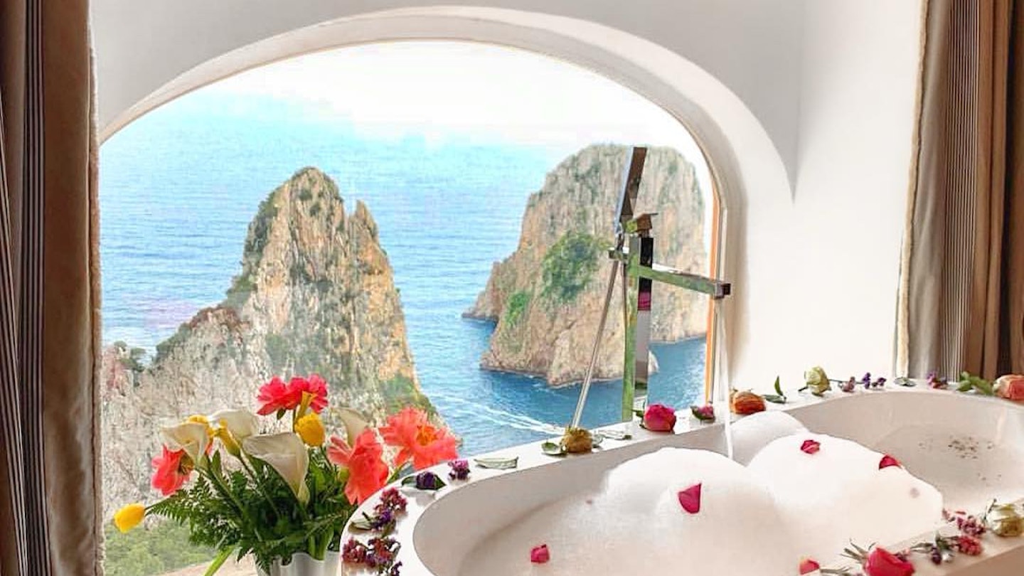 A white soaking tub filled with soap bubbles, with flower petals around the rim, in front of an arched window overlooking the water and rock formations, at Punta Tragara hotel