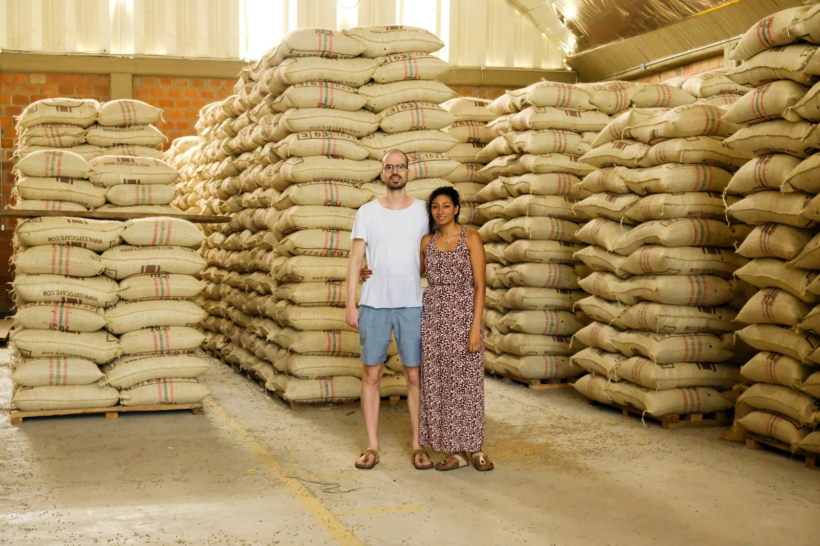The owners of Pacandé coffee roasters in Munich stand in front of hundreds of sacks of coffee piled high on wooden pallets in a large, light-filled red-brick warehouse, smiling at the camera. 