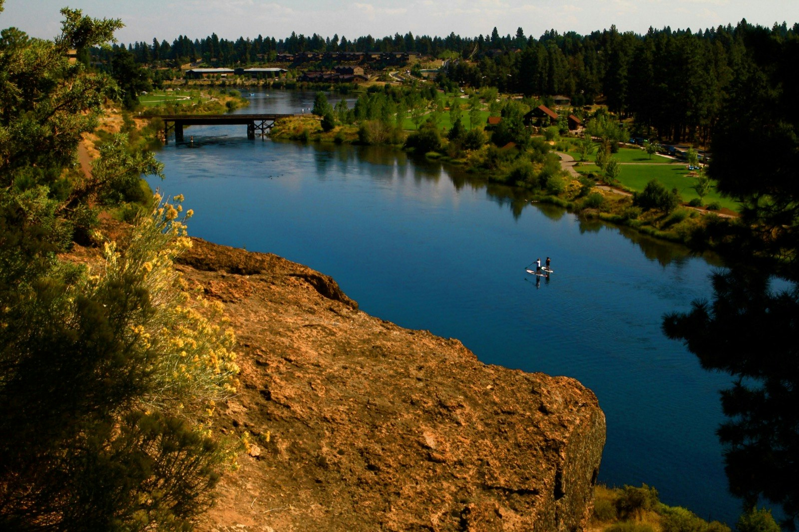 Two people are seen paddleboarding on a deep blue river from a vantage point far above on a rocky outcropping near Bend, Oregon