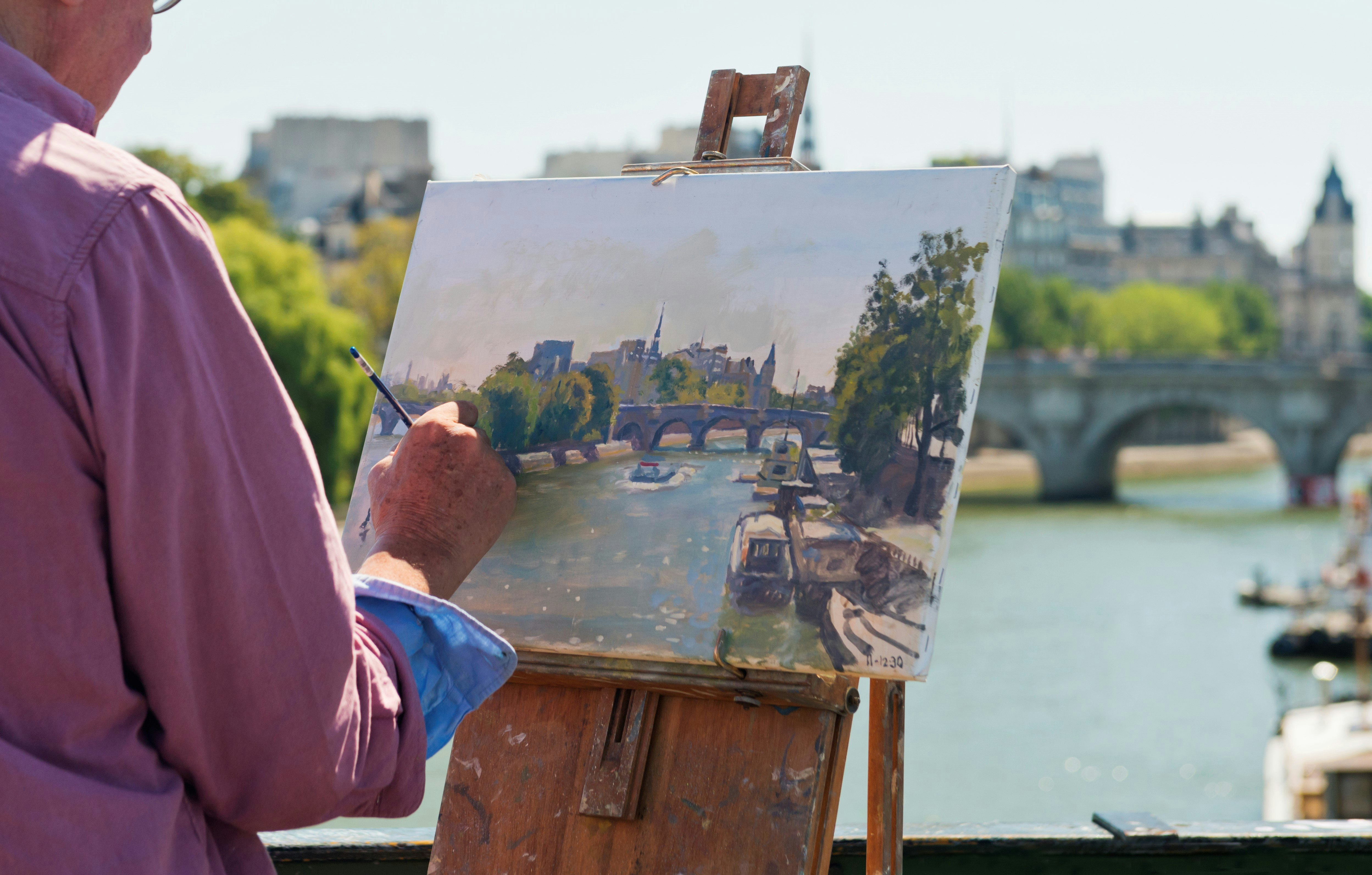 An artist paints a scene of Paris, including the Cathédrale Notre Dame, while standing on a bridge over the River Seine.