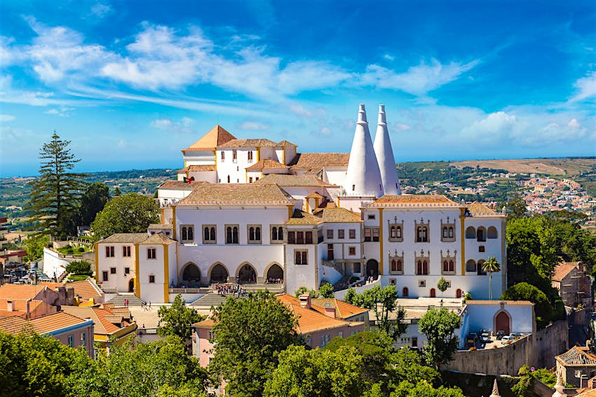 Photo of Palácio Nacional de Sintra (Sintra National Palace); the large complex sits on top of a hill and has whitewashed walls, a terracotta-tiled roof, and two conical white turrets.