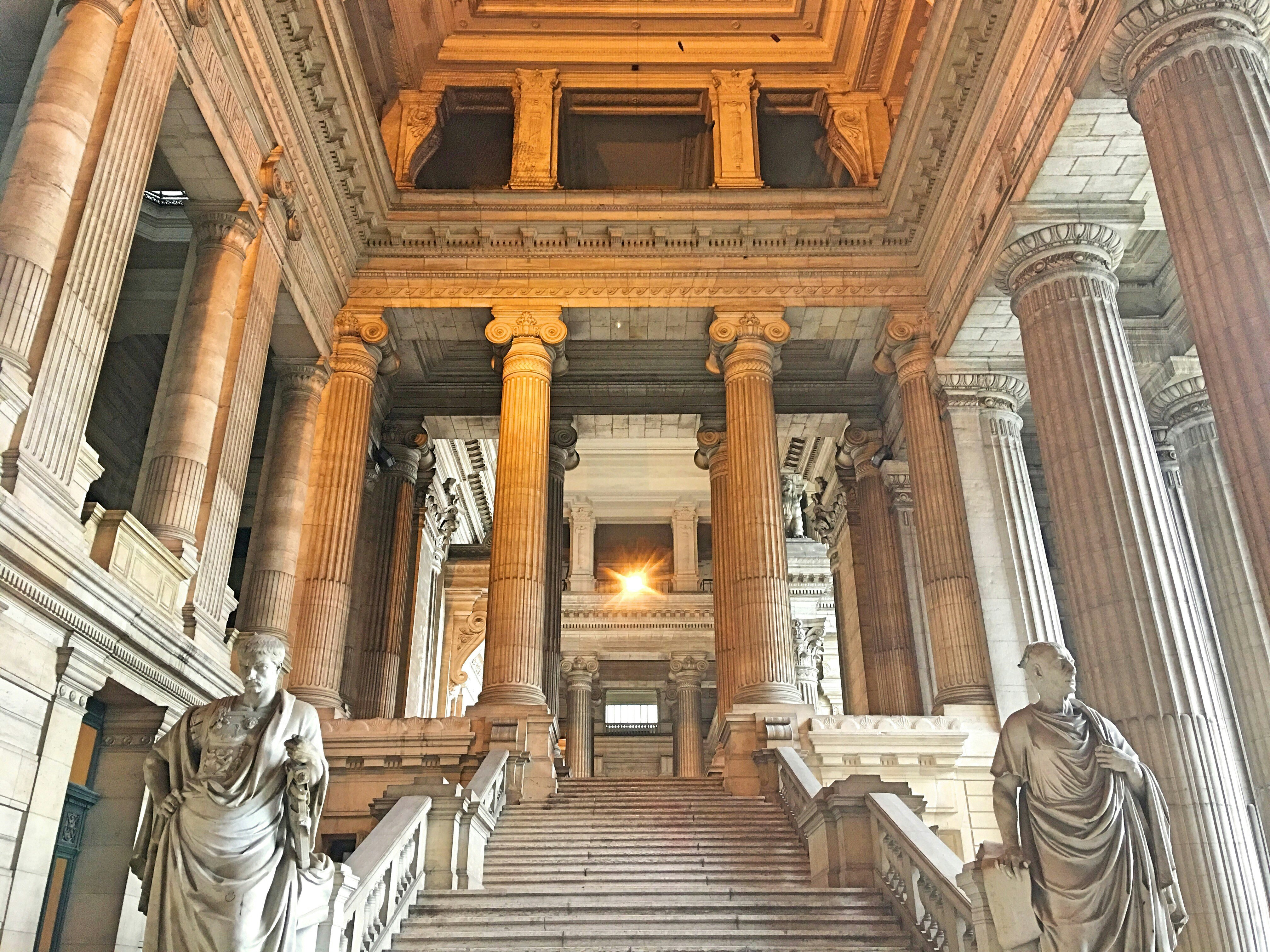 Stone steps flanked by statues leading up to the grand, galleried and colonnaded Palais de Justice interior.