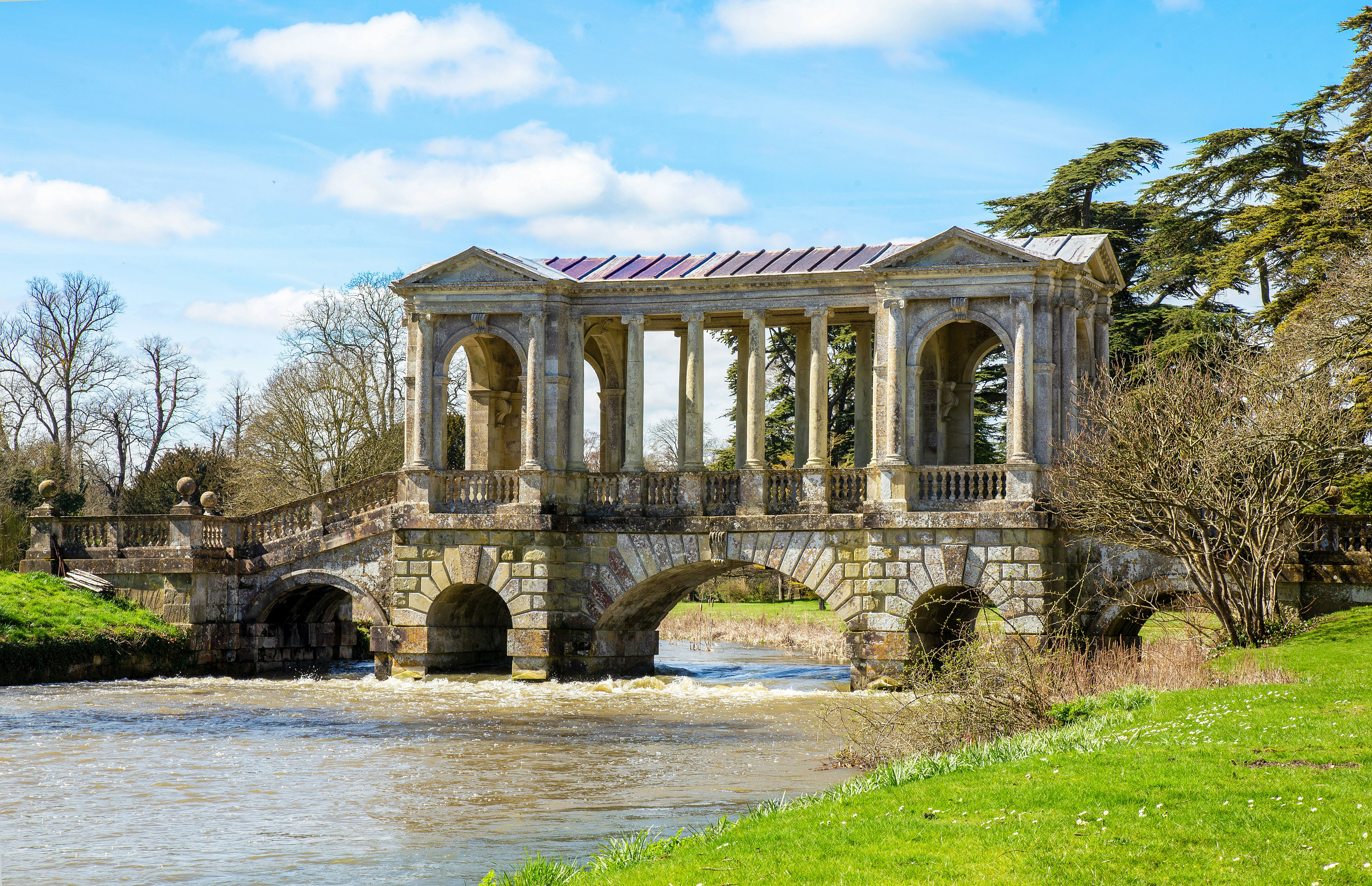 A stream flows beneath the Palladian Bridge at Wilton House. Trees without their leaves are visible in the background.