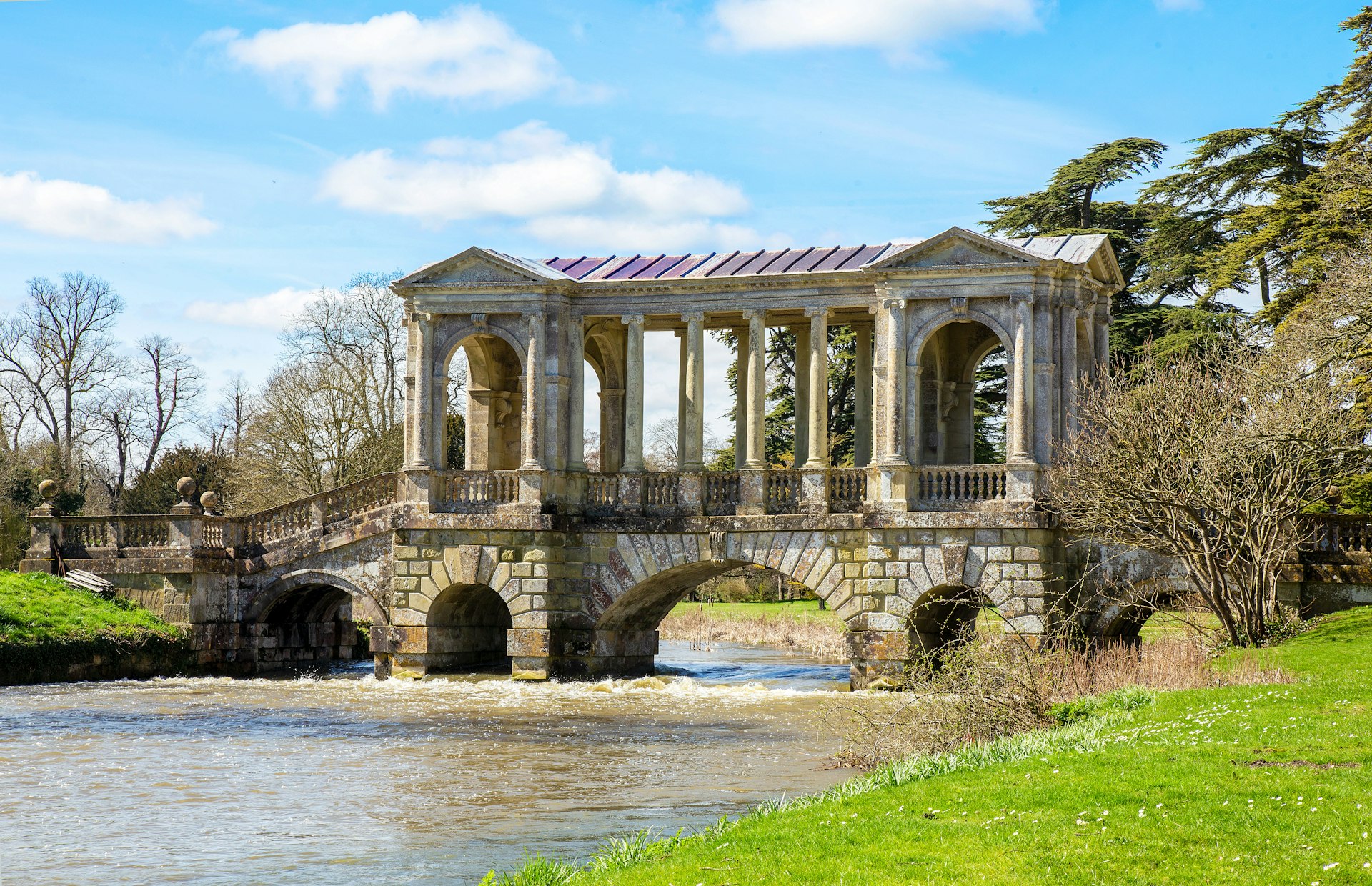 A stream flows beneath the Palladian Bridge at Wilton House. Trees without their leaves are visible in the background.