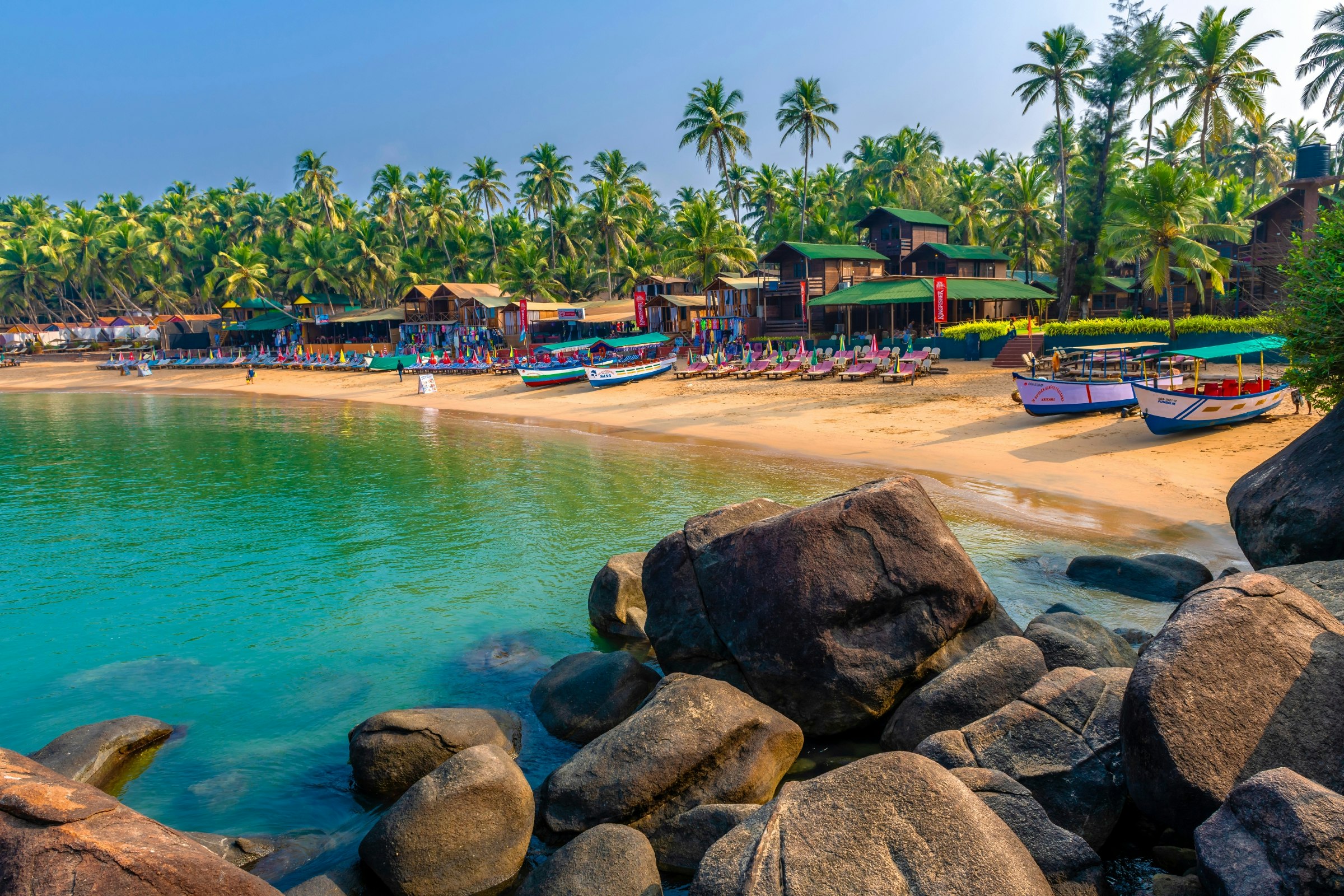 A row of colourful bungalows line the sands of Palolem in Goa. The sea is calm and blue, and behind the beach, palm trees are visible. A few people sit on the sand.