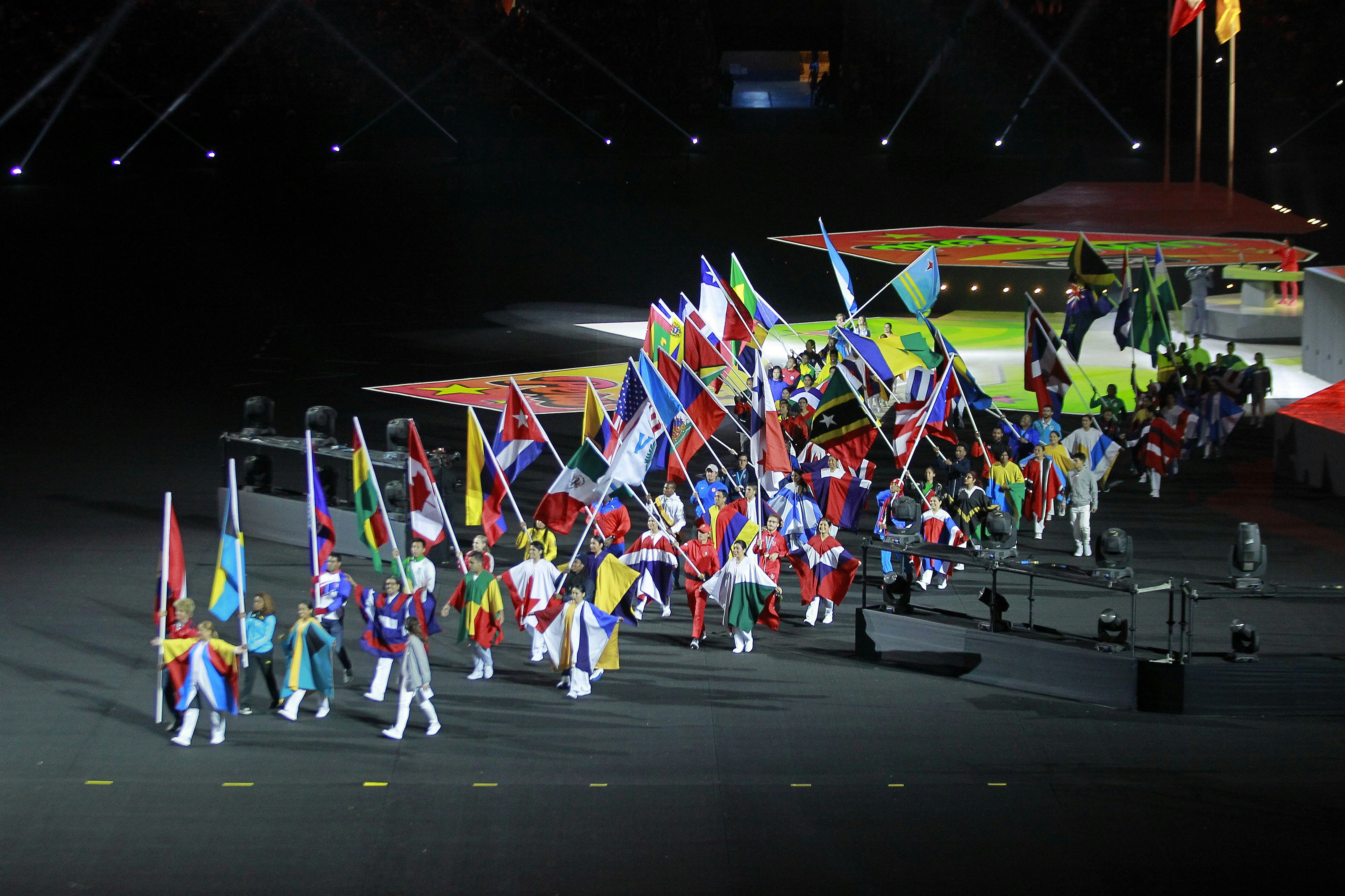 A group of people dressed in and holding national flags walk across a stage during the 2019 Pan American Games in Lima
