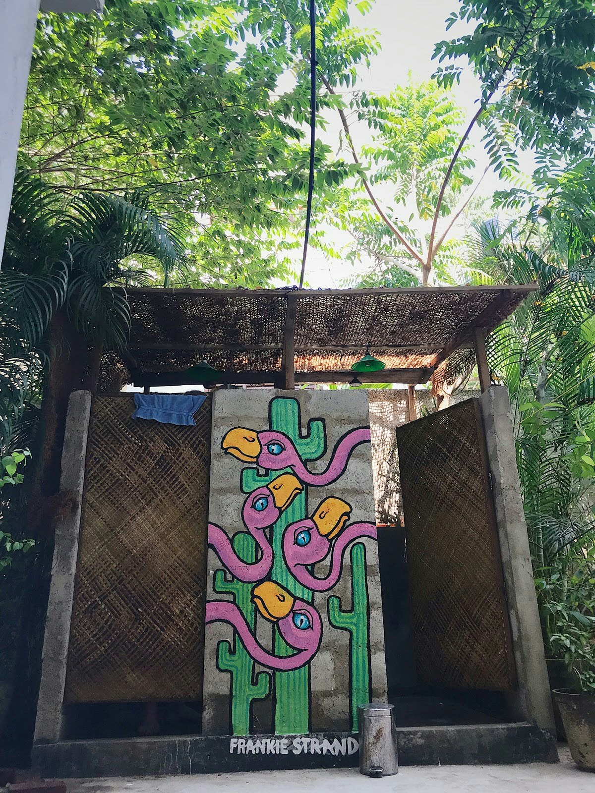 A mural painted on the wall of two outdoor showers. The shower cubicles have woven wicker doors and sit under a canopy of tropical leaves with blue sky peeking through. The colourful mural shows four flamingo heads set against a backdrop of cacti.