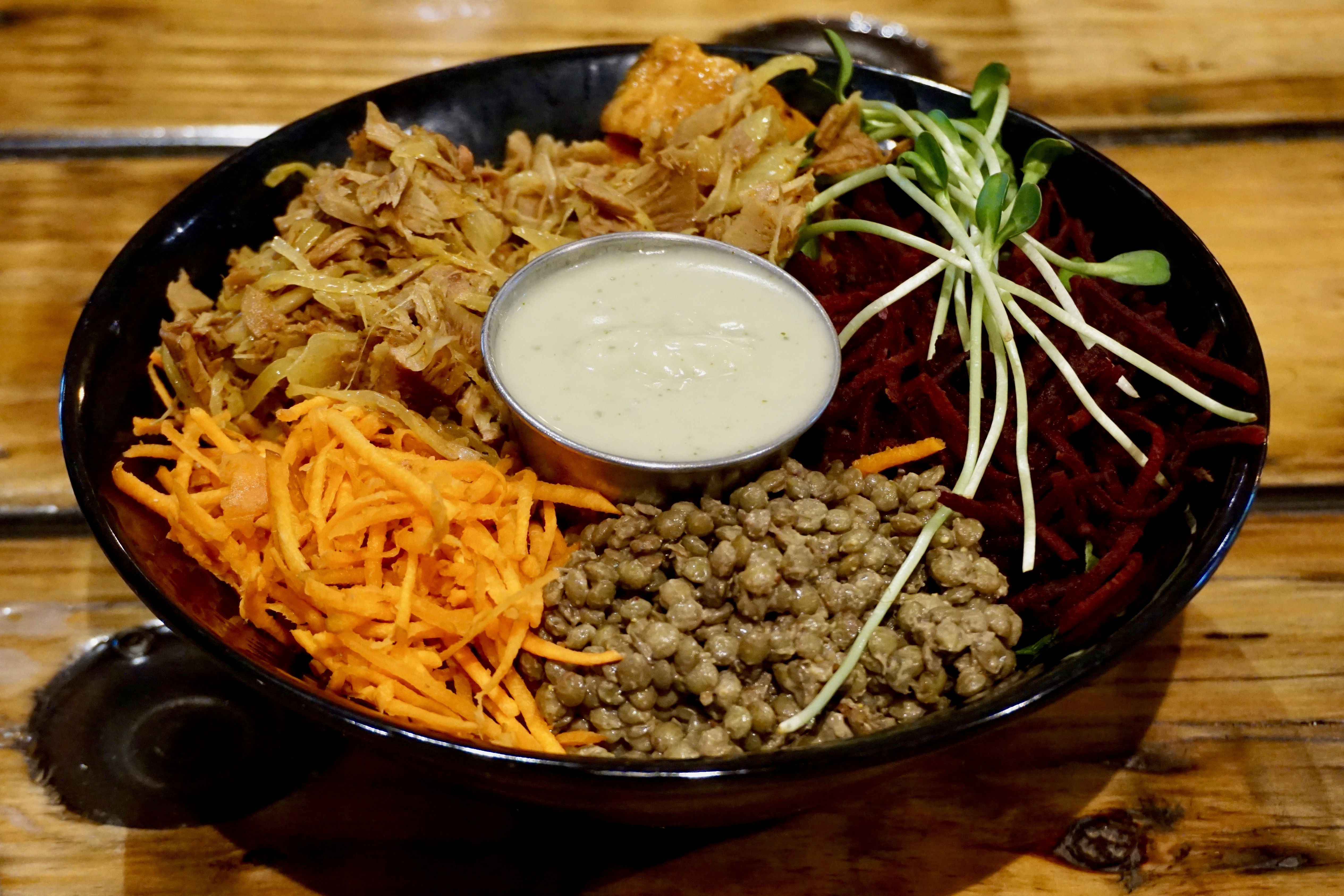 A large black bowl is filled with pulled jackfruit, grated beets and carrots and bulgar wheat. In the middle is a stainless steel ramekin with a white sauce.