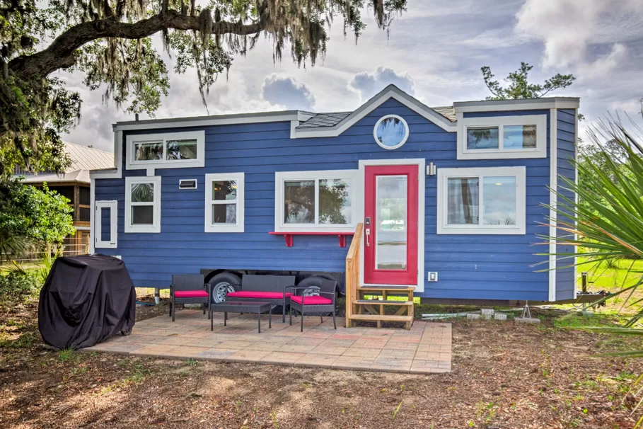A tiny house on wheels that's painted blue and has seats in front of it