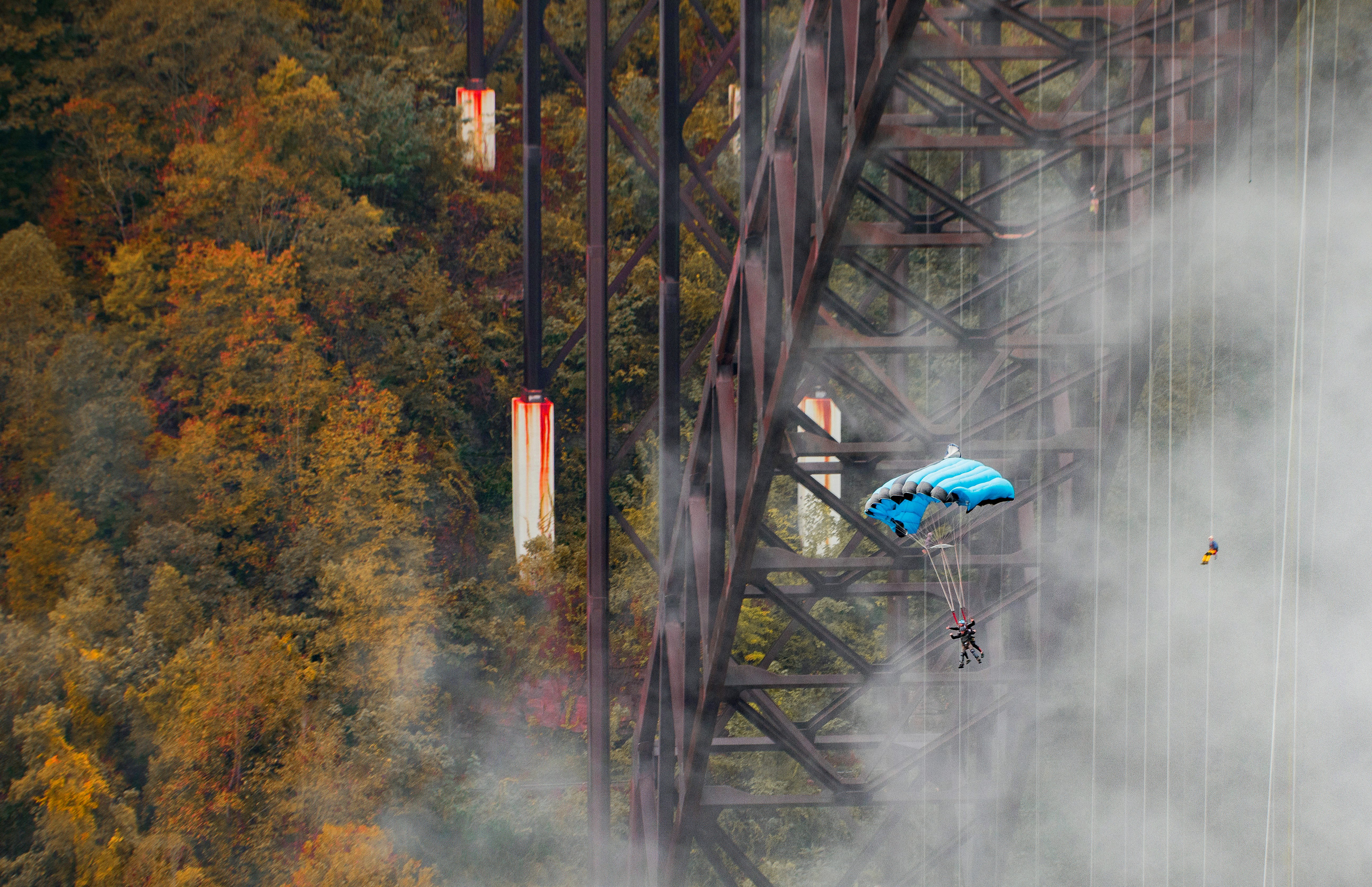 Two people attached to a parachute are gliding under New Gorge Bridge during the day. Woodland and support beams for the bridge are visible.