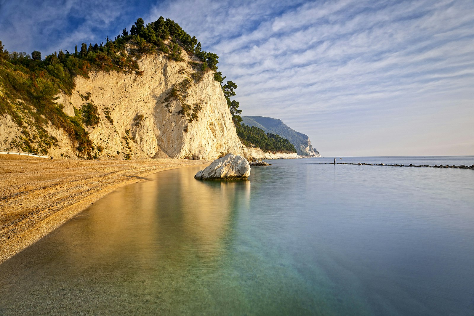 A view from the water of the rocky coastline of Parco del Conero; the cliff face is white and topped with evergreen trees, and a golden stretch of sand runs up to the hill. Le Marche, Italy.