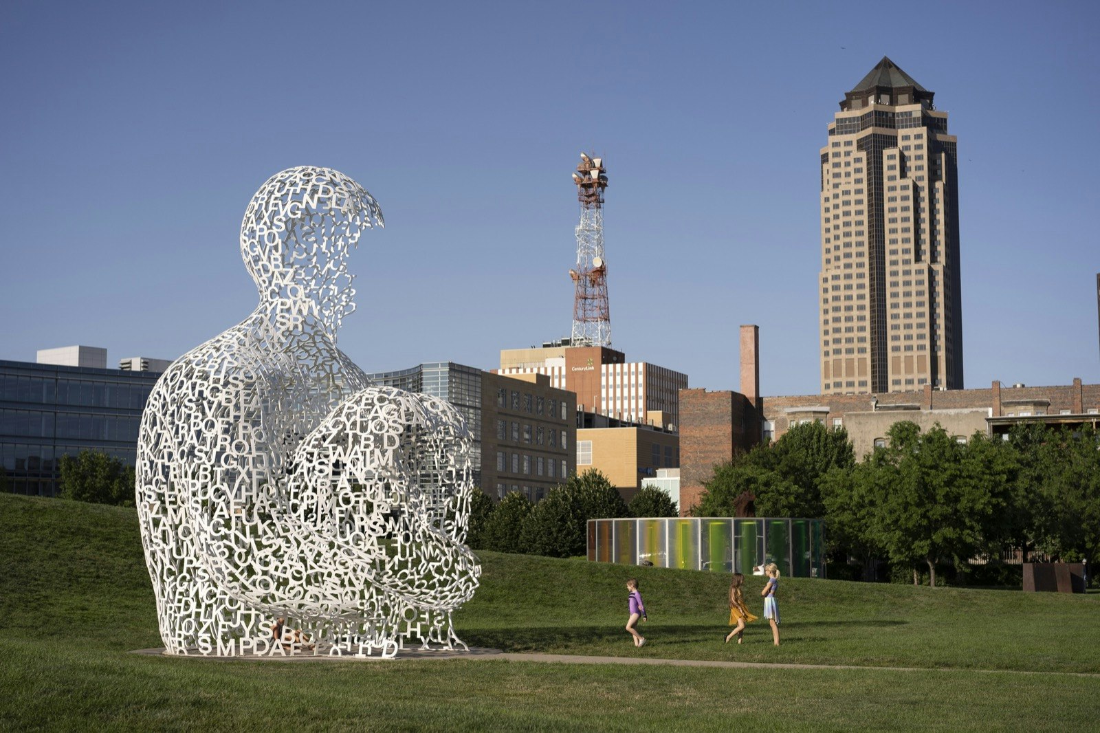 A massive sculpture of a man, made of white letters of the alphabet, sits in a park surrounded by a tall building and radio tower; Things to do in Des Moines during the Iowa Caucuses