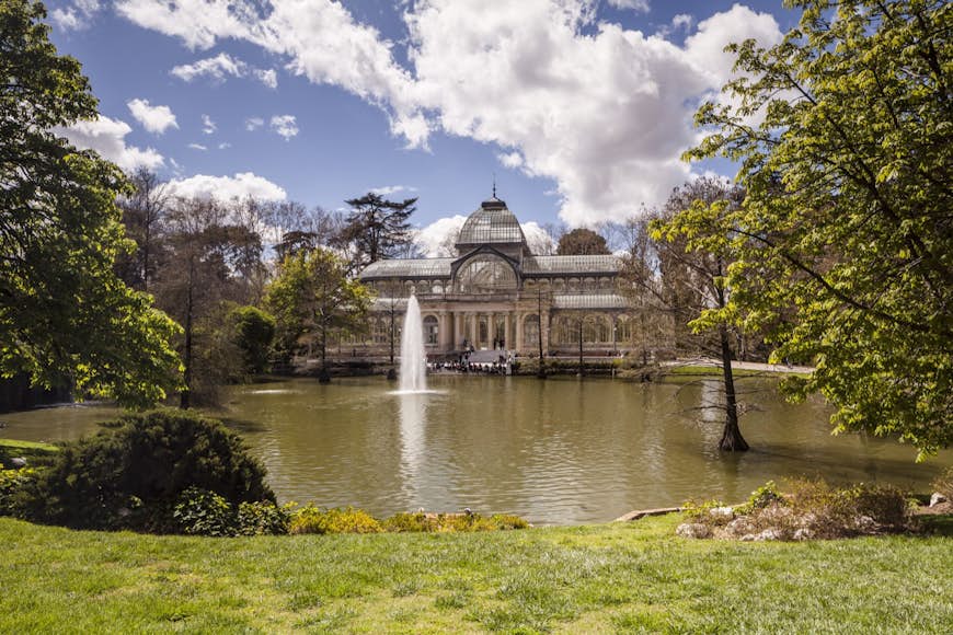 An arched glass and iron building in front of a lake with a fountain in the middle