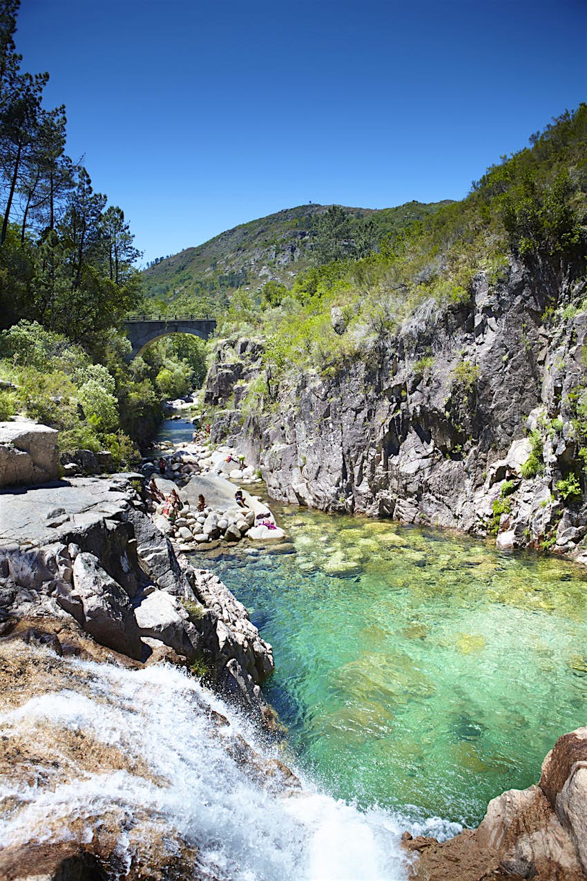 A crystal clear pool of turquoise water sits within a narrow rocky gorge, with a forested hill in the background.