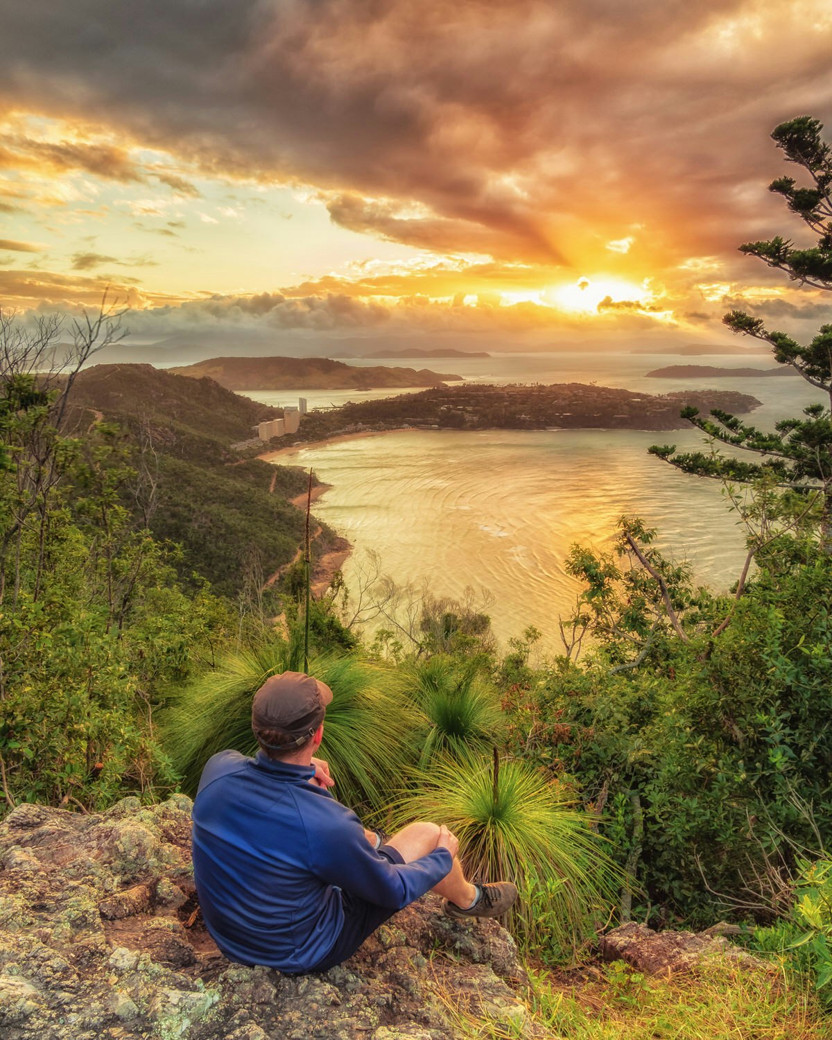 A man sits on a rock at Passage Peak looking at a sunset over the sea on Hamilton Island.