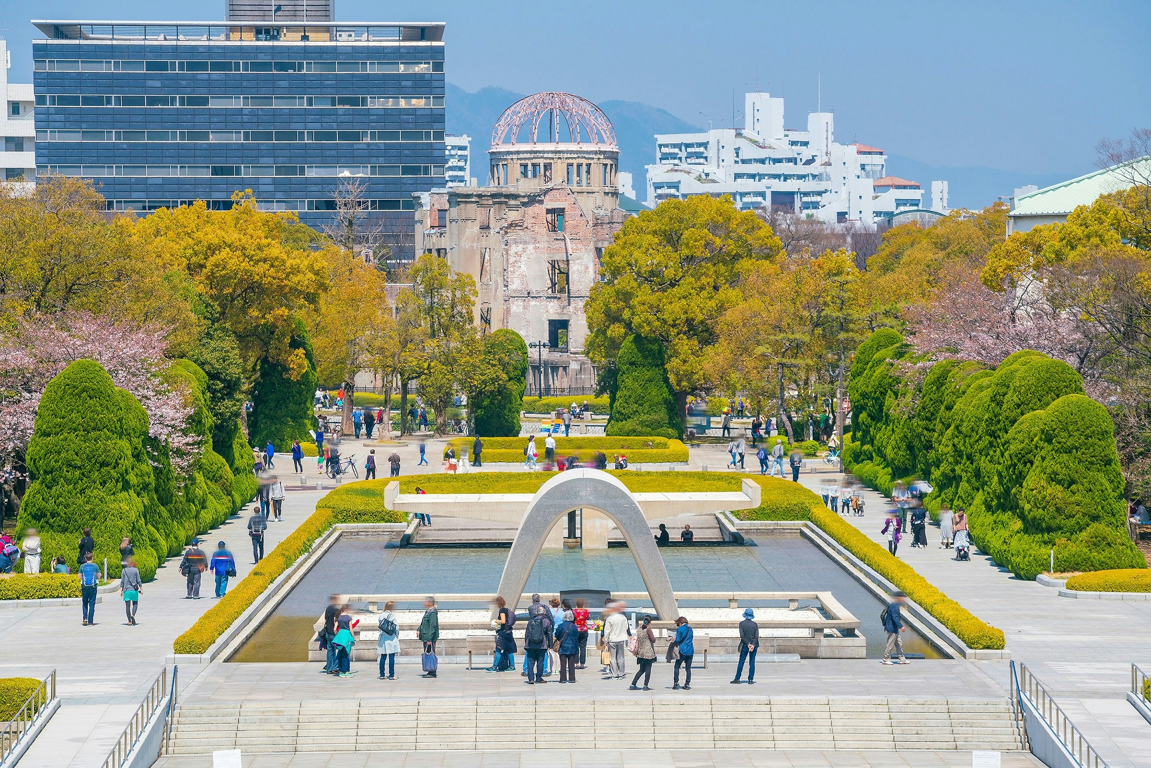 An aerial view overlooking the Peace Memorial Park in Hiroshima. The park has a small pond in the centre of it, with trees and lawns all around it. Many people wander along the pathways flanking the pond.
