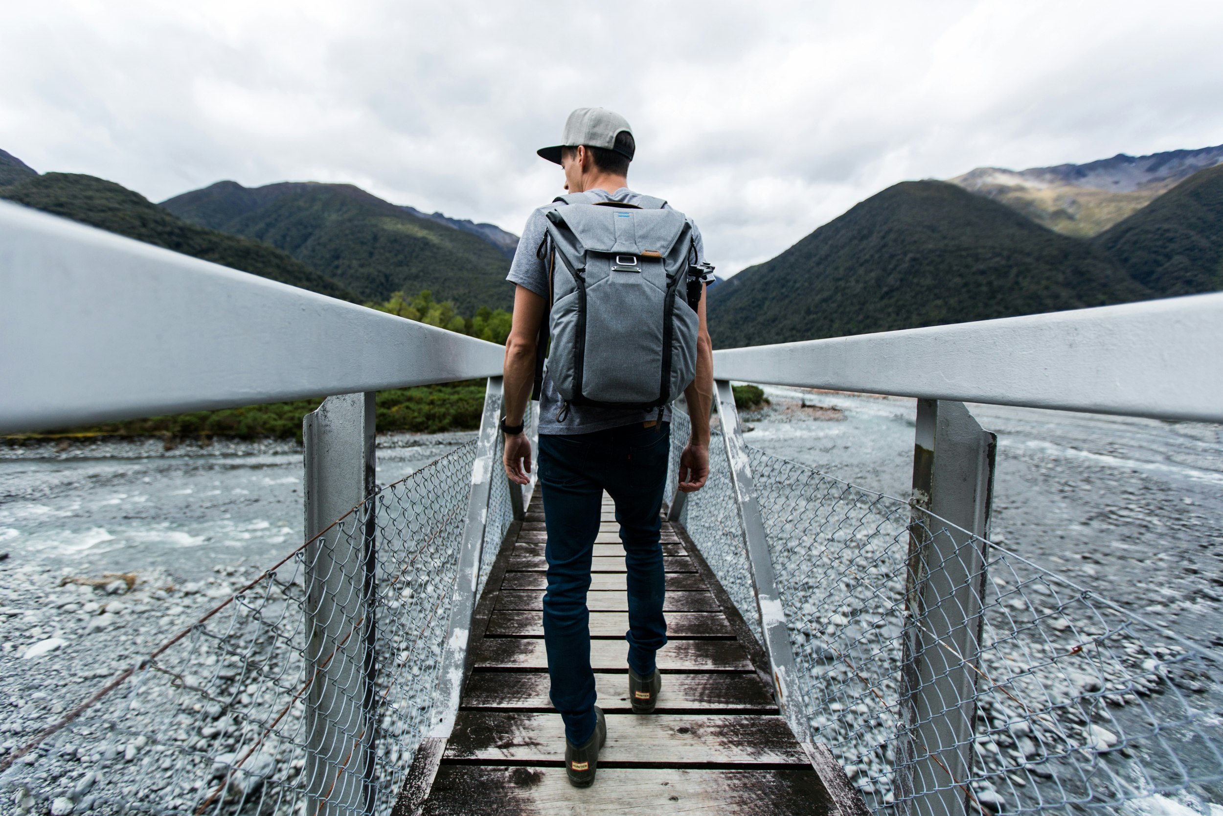 A man wearing an Everyday backpack is going across a walkway with wooden rails. The ground either side of him is covered in gravel, mountains are in the near distance. 