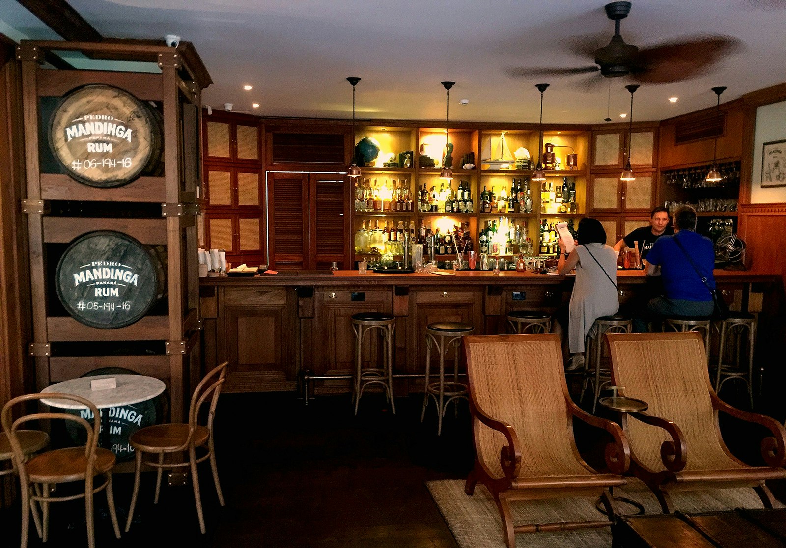 A man and a woman sit at at wooden bar in front of a bartender. The woman is holding a menu. To the left is a shelf filled with large wooden barrels. In front of the shelves is a small table with a pair of chairs. There are two rattan campaign chairs in the middle of the bar. 