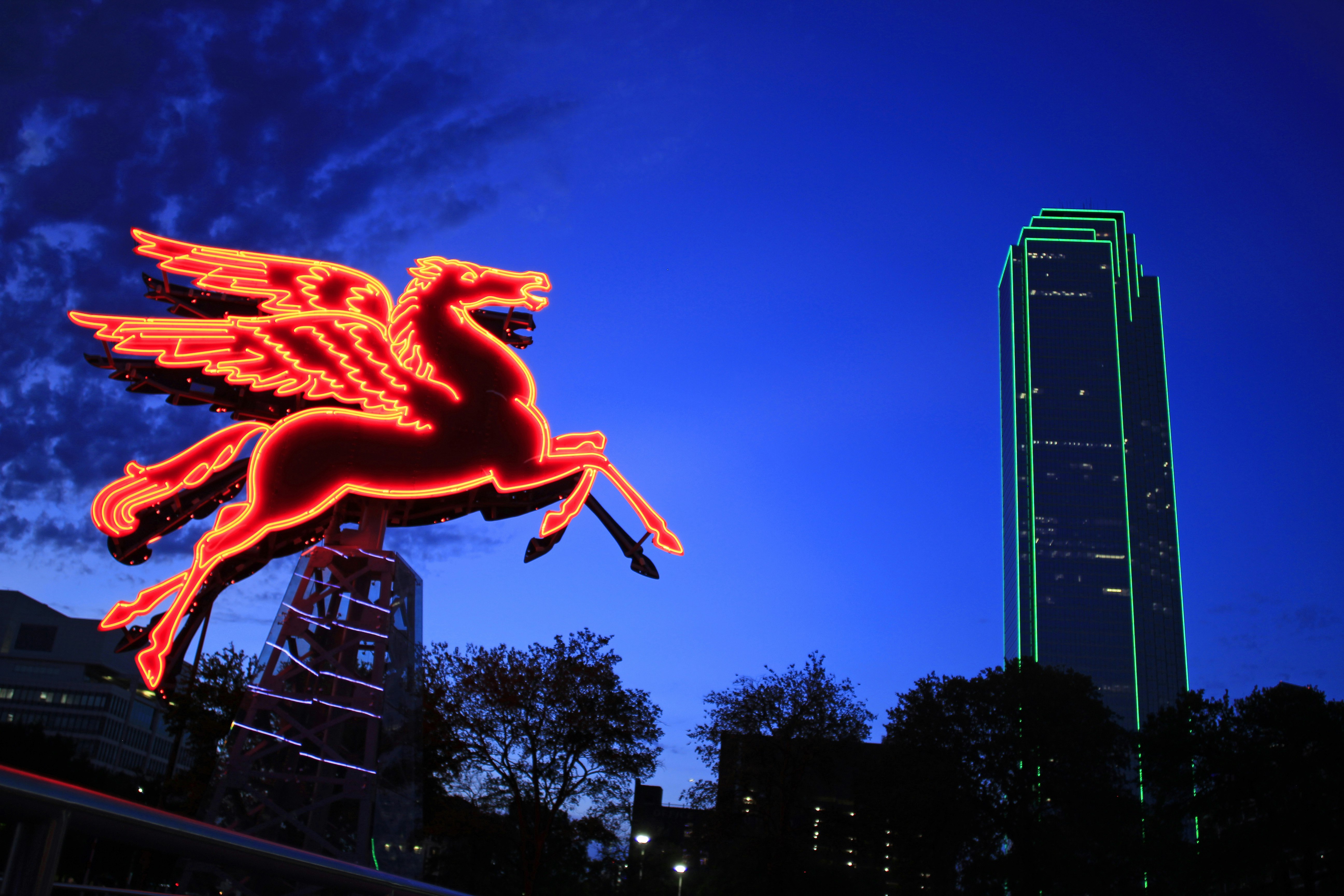 A neon sign of a Pegasus glows in the evening next to a tall building outlined in neon green lights; Thanksgiving NFL city