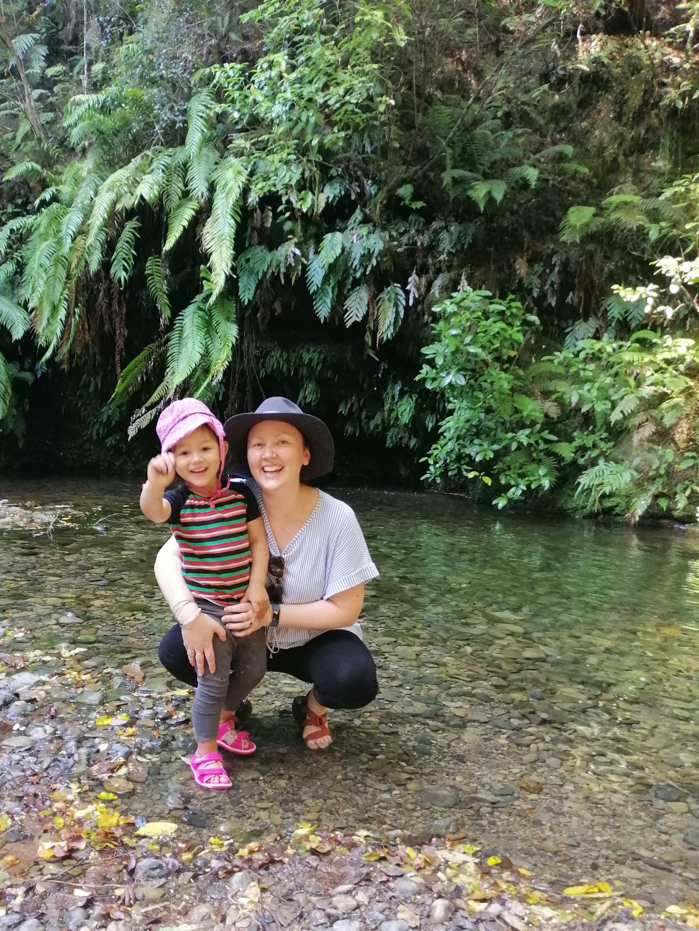 A woman and her daughter are standing on rocky ground and smiling towards the camera. Both are wearing hats.