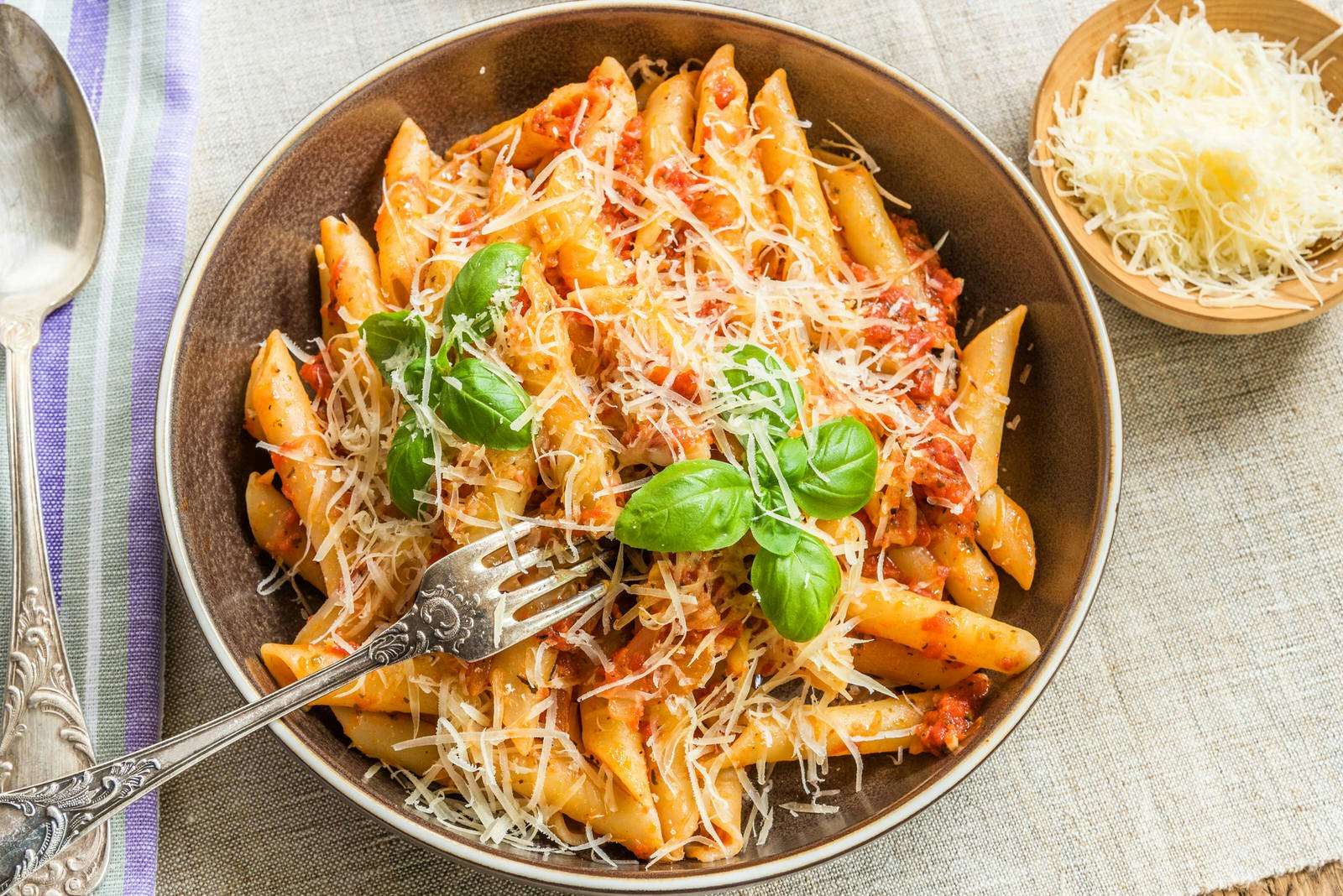 Pasta All'Arrabbiata, penne with tomato sauce and basil and parmesan cheese on a rustic wooden table with linen napkin