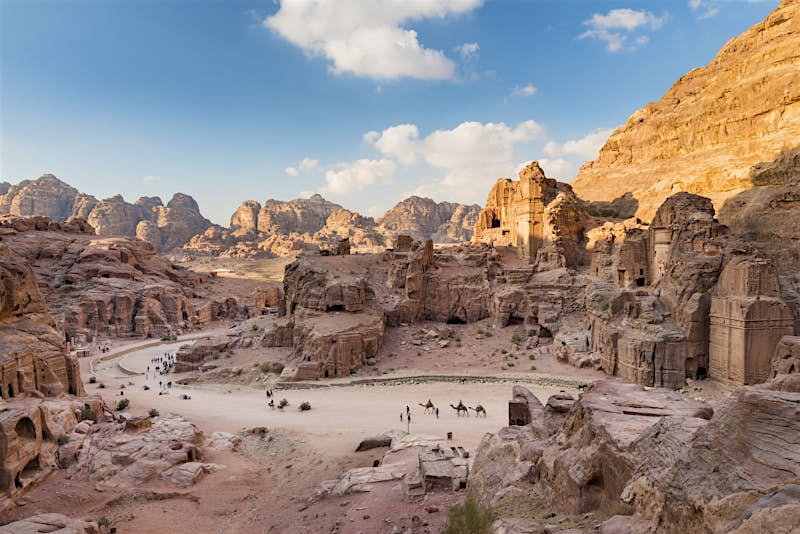 A distant shot of the Petra site. Structures are cut out of the red rocks and there are mountains in the distance.
