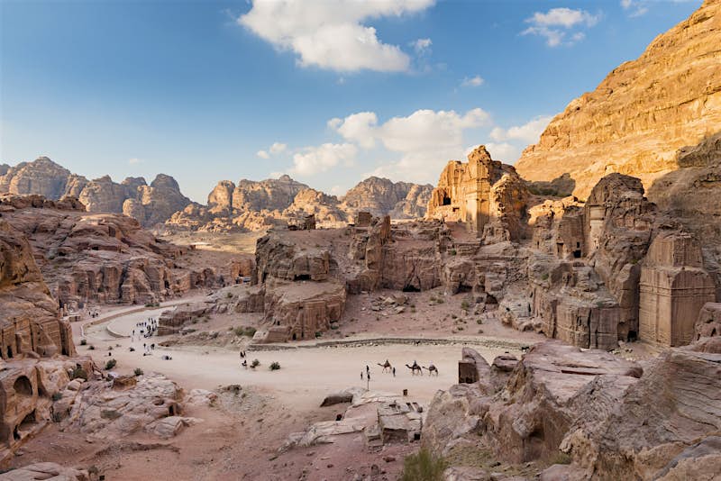 Petra in Jordan A distant shot of the Petra site. Structures are cut out of the red rocks and there are mountains in the distance.