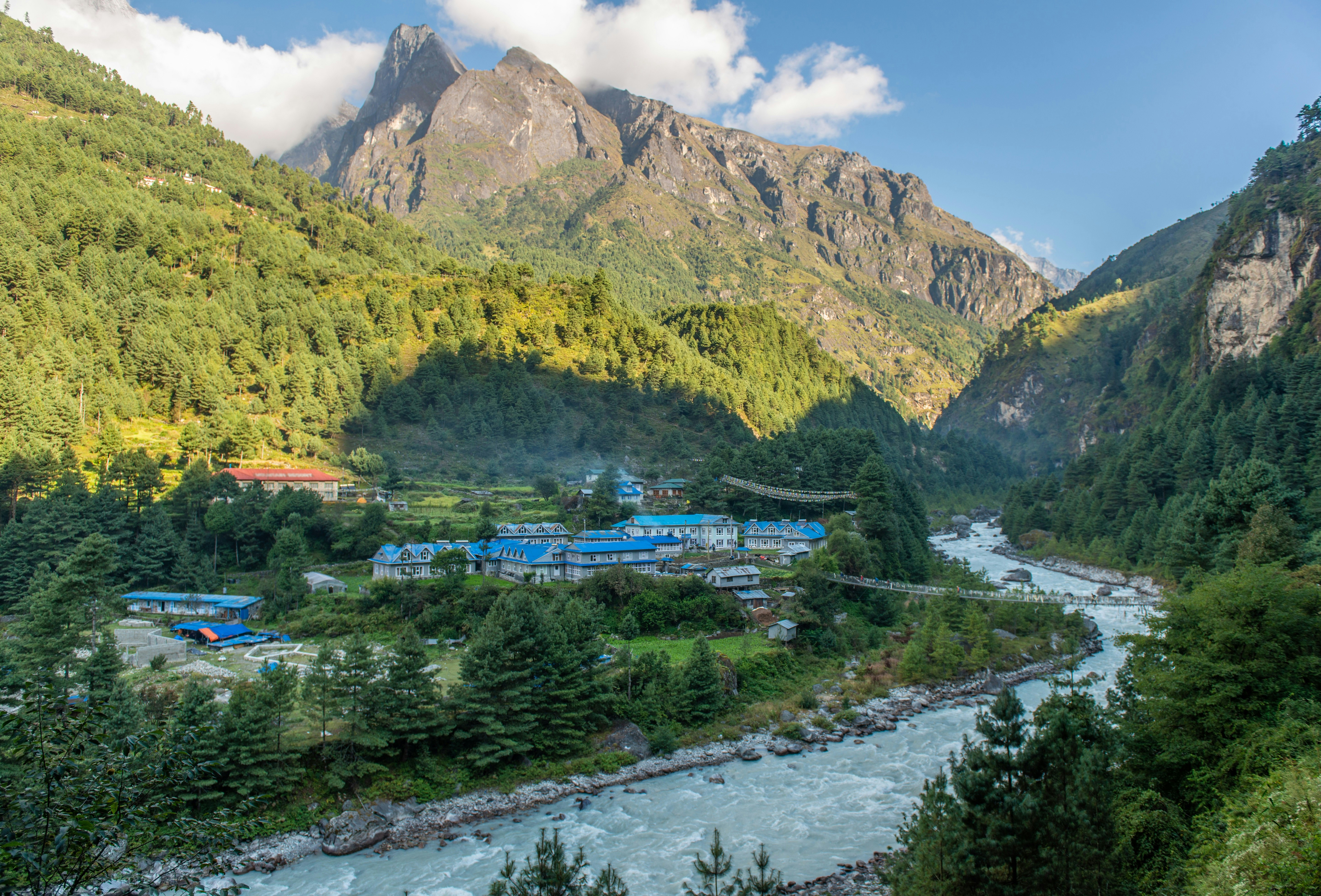 A cluster of blue-roofed buildings that make up the small town of Phakding, a stop on the trekking route to Everest Base Camp. A lovely blue river sweeps around the town, while the surrounding hills are covered in forest.