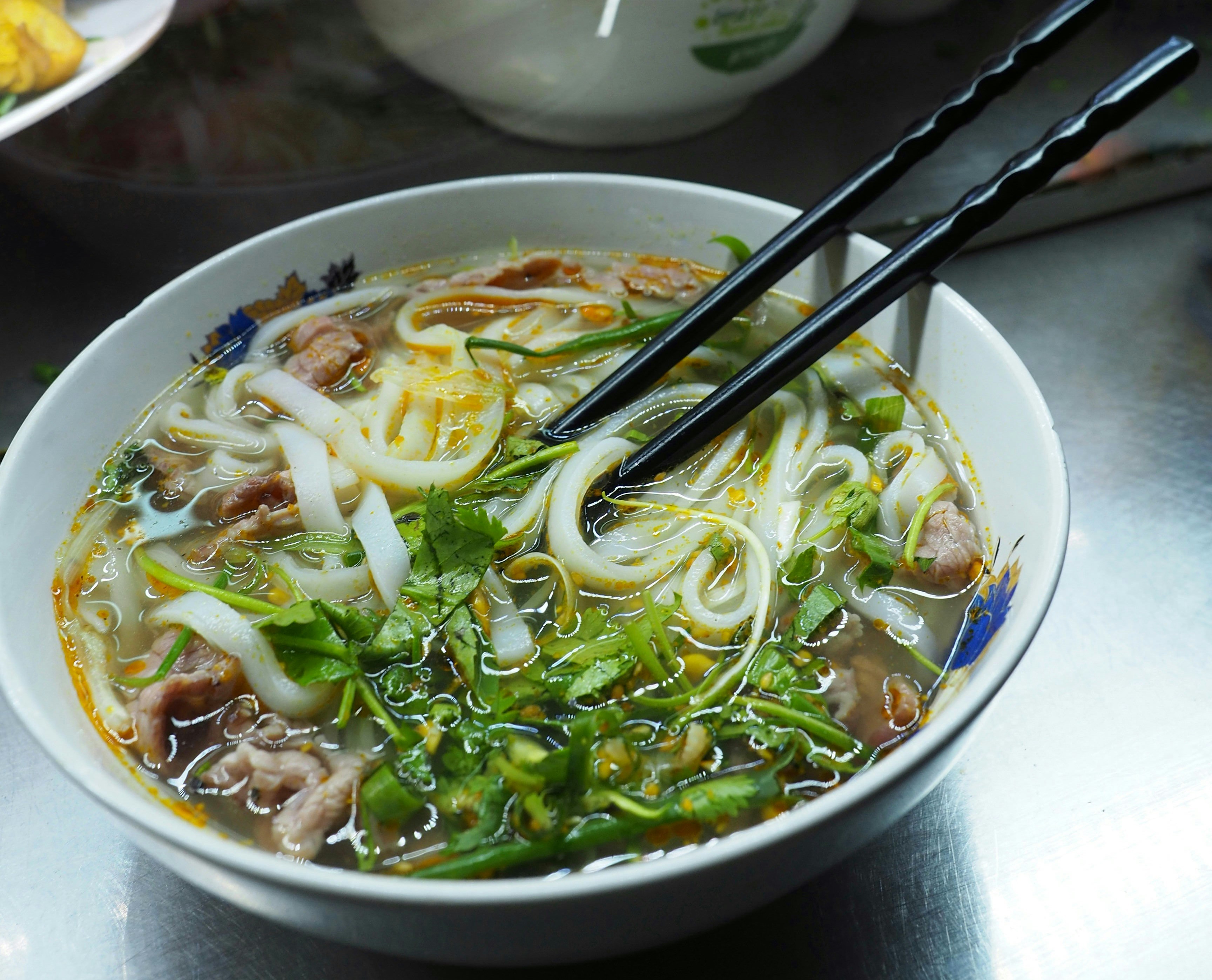 A side-on view of a bowl of pho stood on a tabletop in a Vietnamese restaurant. The pho contains thick noodles, veg and meat, and has a pair of chopsticks resting in it.