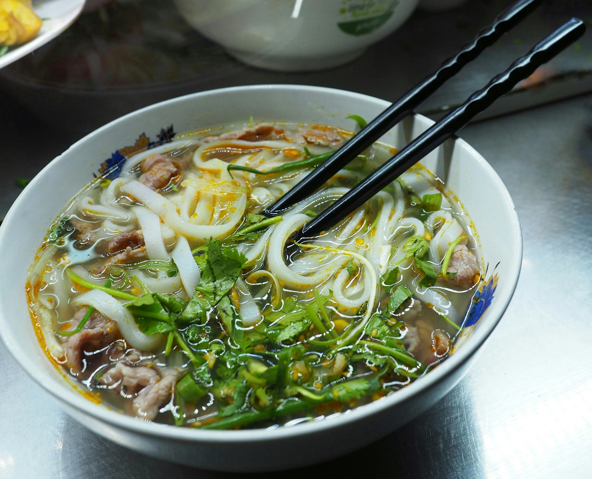 En sidovy av en skål med pho stod på en bordsskiva på en vietnamesisk restaurang.  Pho innehåller tjocka nudlar, grönsaker och kött, och har ett par ätpinnar vilande i den.