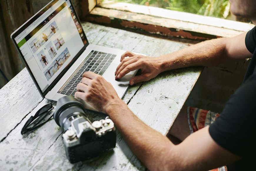 En man sitter på ett slitet bord och redigerar bilder på en bärbar dator.  Hans kamera sitter bredvid den bärbara datorn på skrivbordet. 