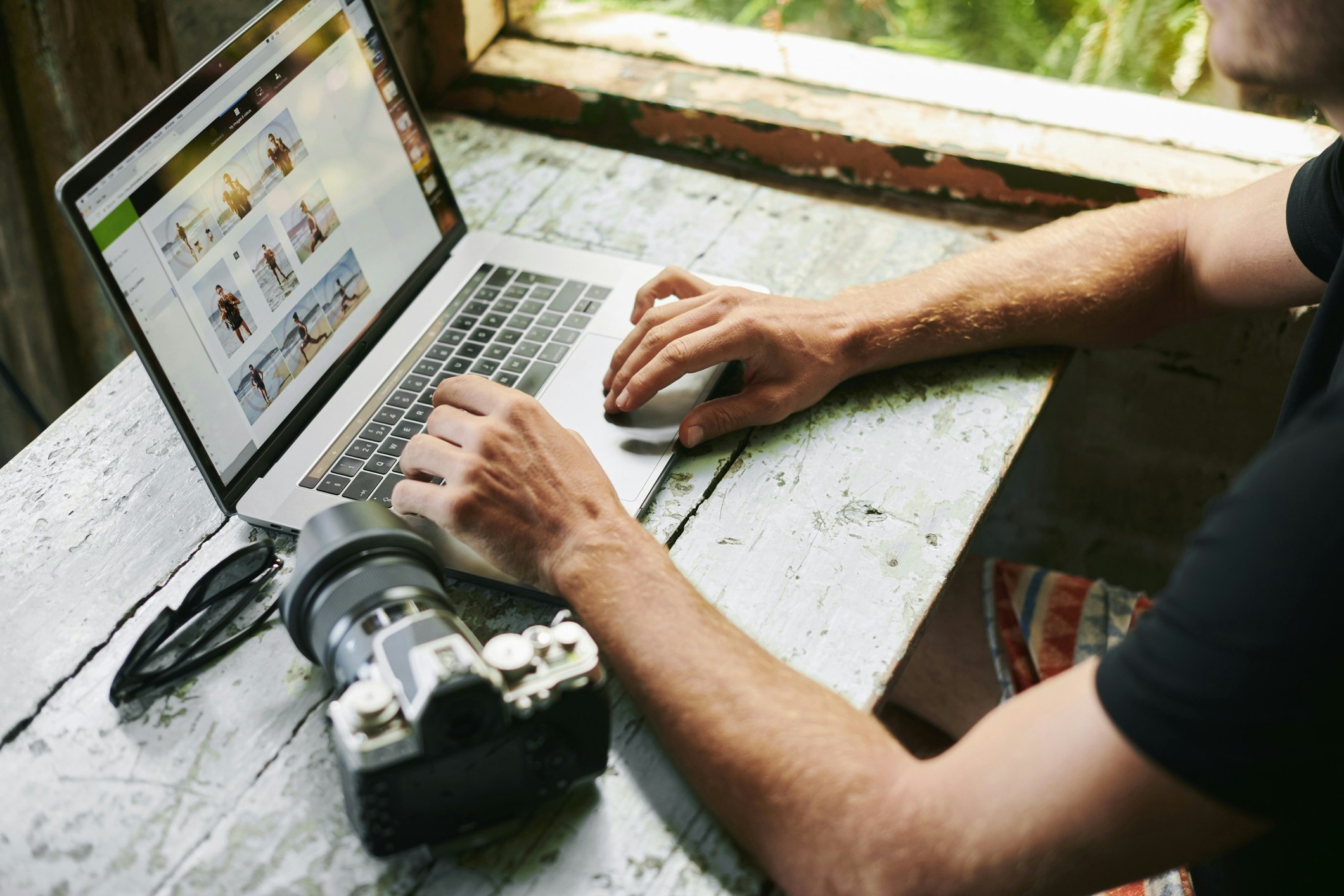 A man is sitting a worn-out table editing photos on a laptop. His camera is sitting next to the laptop on the desk. 