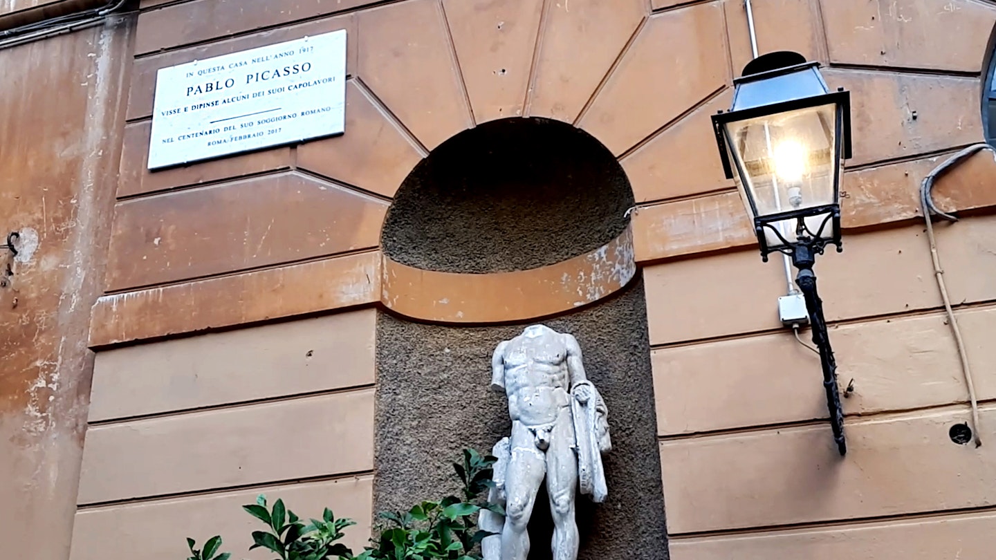 A sign marks the home of Pablo Picasso, positioned on the building above and to the left of an alcove with a statue of a headless nude statue 