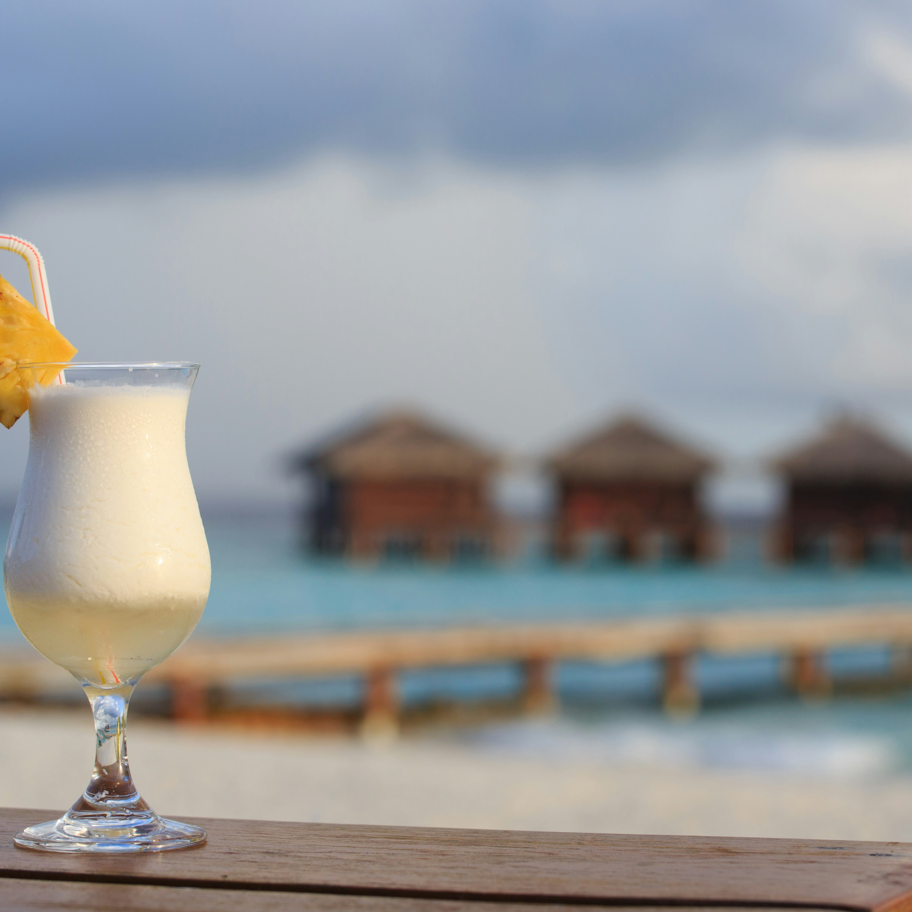 A poco grande glass of Pina Colada garnished with a pineapple wedge and a red and white stripped drinking straw sits on a wooden ledge. In the background you can see four bungalows in the ocean.