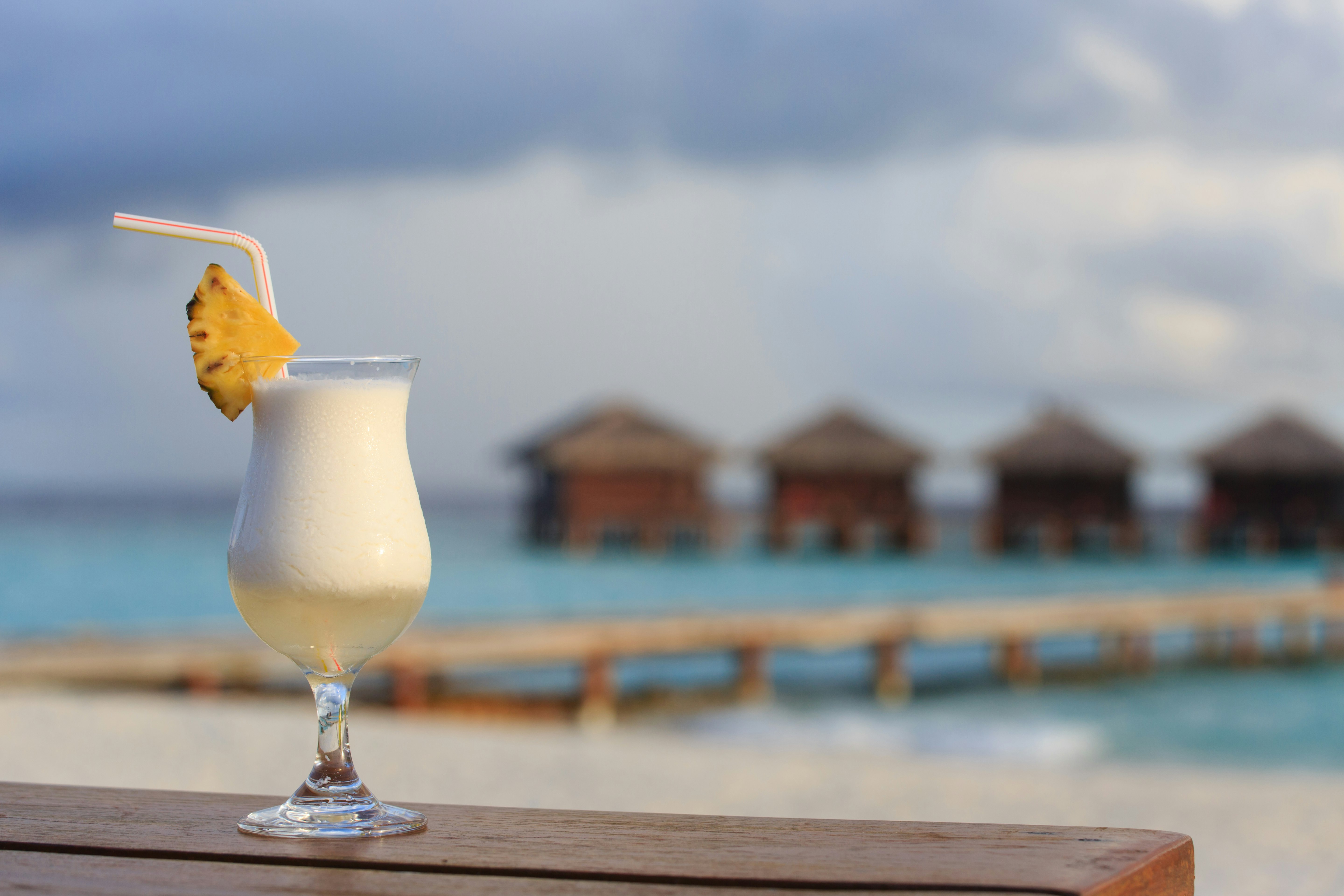 A poco grande glass of Piña Colada garnished with a pineapple wedge and a red and white stripped drinking straw sits on a wooden ledge. In the background you can see four bungalows in the ocean.