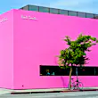 A building on the corner of a street has bright pink walls. A man stands nearby with a small dog on a lead
