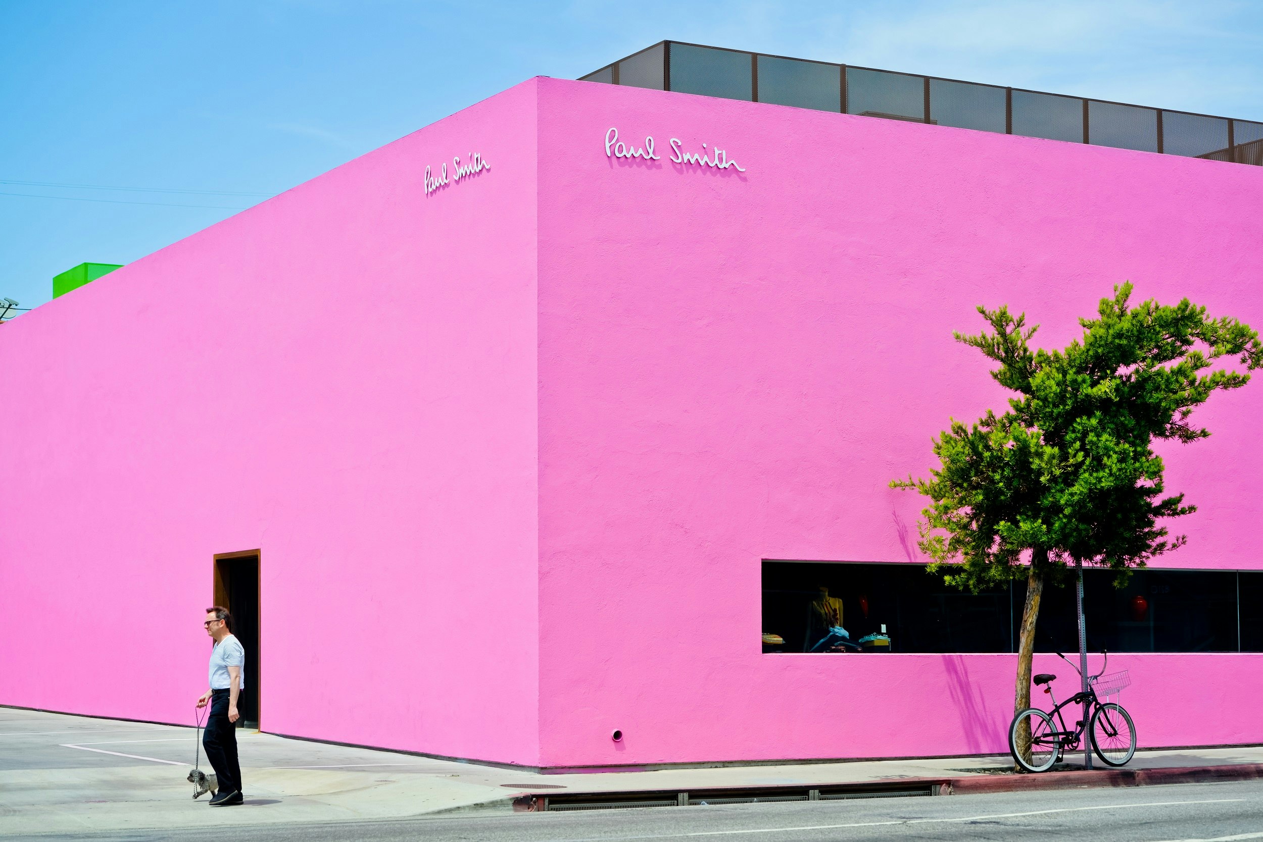 A building on the corner of a street has bright pink walls. A man stands nearby with a small dog on a lead