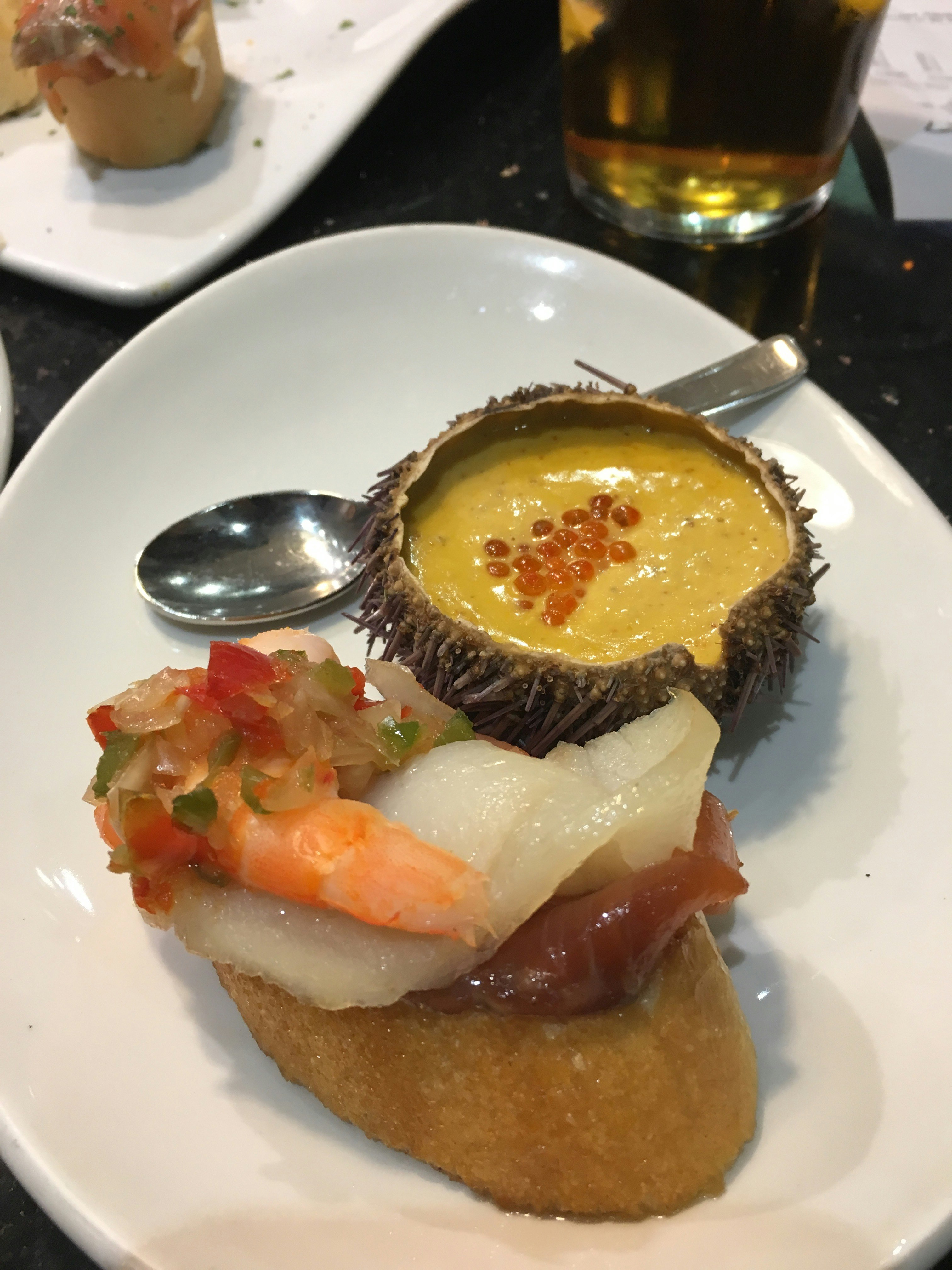 A creamy yellow soup is served inside a sea urchin husk and sprinkled with orange caviar. It sits next to a fried potato wedge topped with shrimp, raw fish, and marinated peppers – an example of pintxos in San Sebastian
